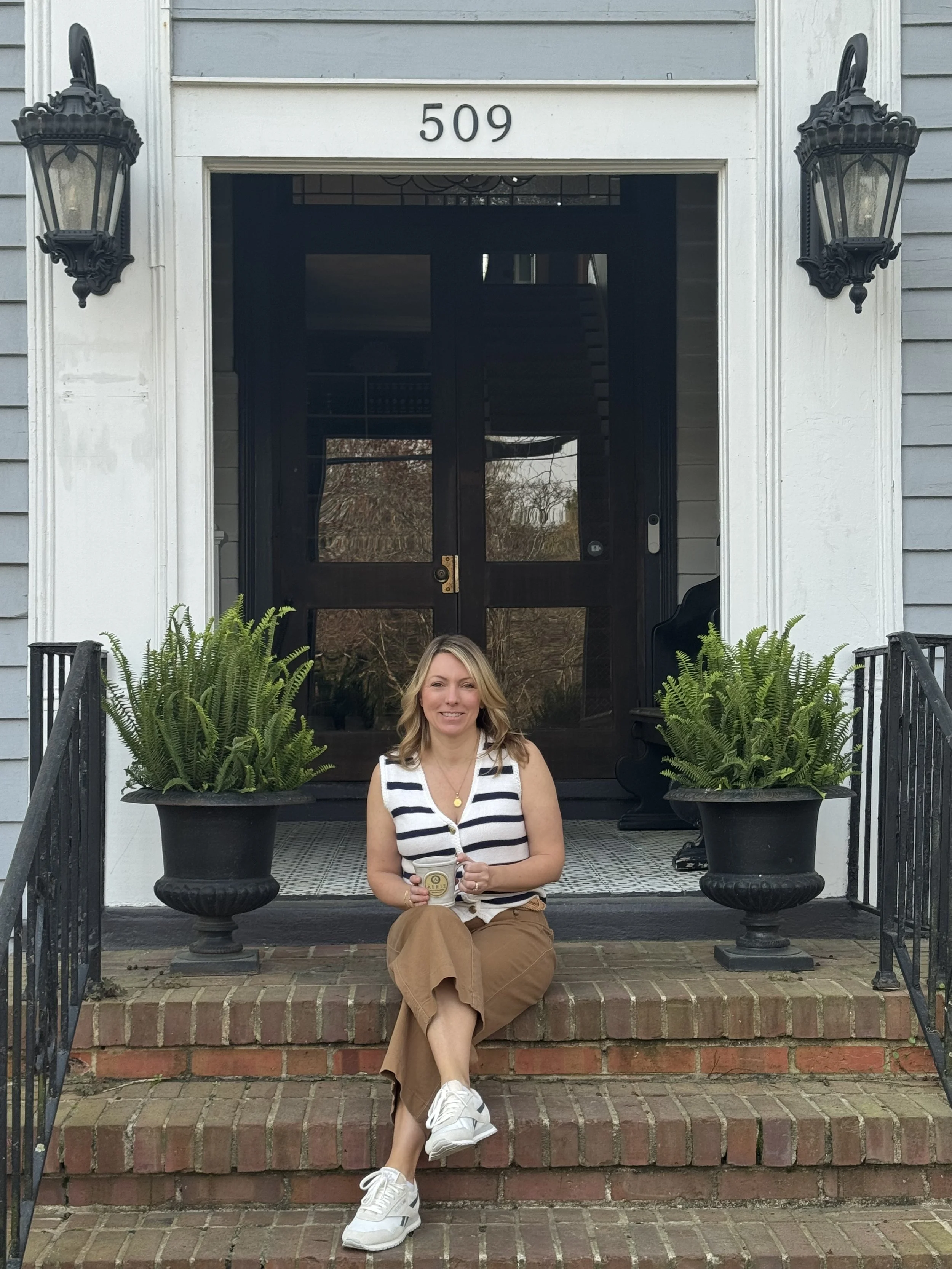 Woman sitting on brick steps outside house with the number 509, holding a drink, wearing a striped top, brown pants, and white sneakers, flanked by large potted plants with ferns, and black lantern-style wall lights.