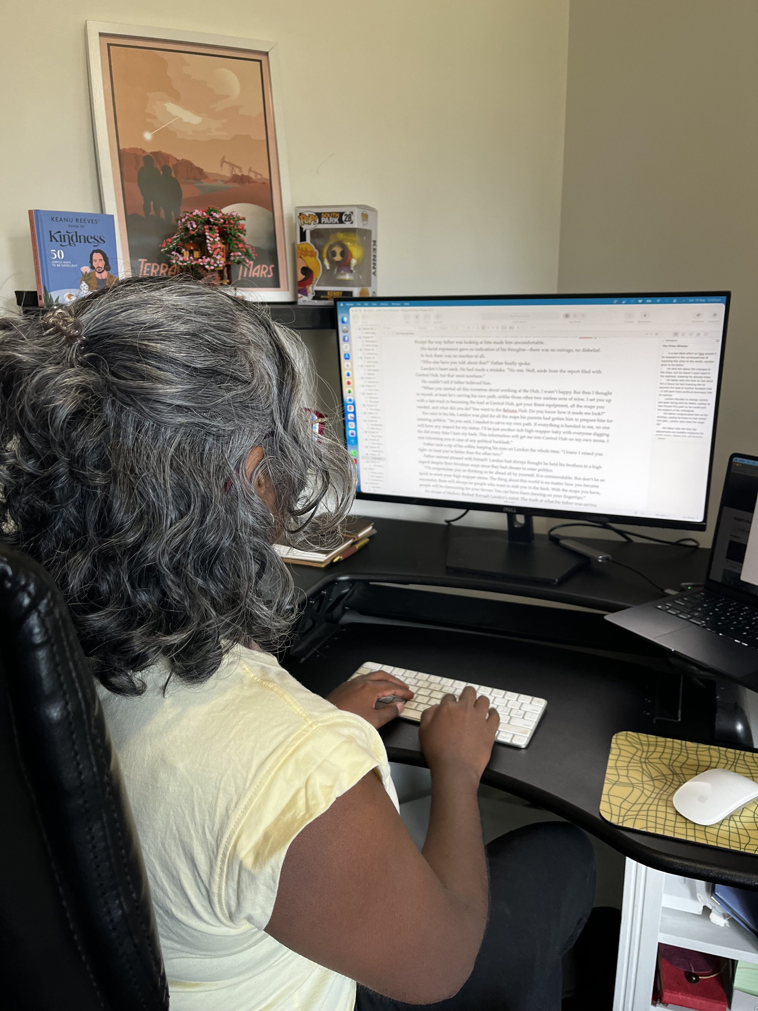 A person with gray curly hair sitting at a desk working on a computer with a large monitor, using a small keyboard. On the wall in the background there are framed posters and figurines.