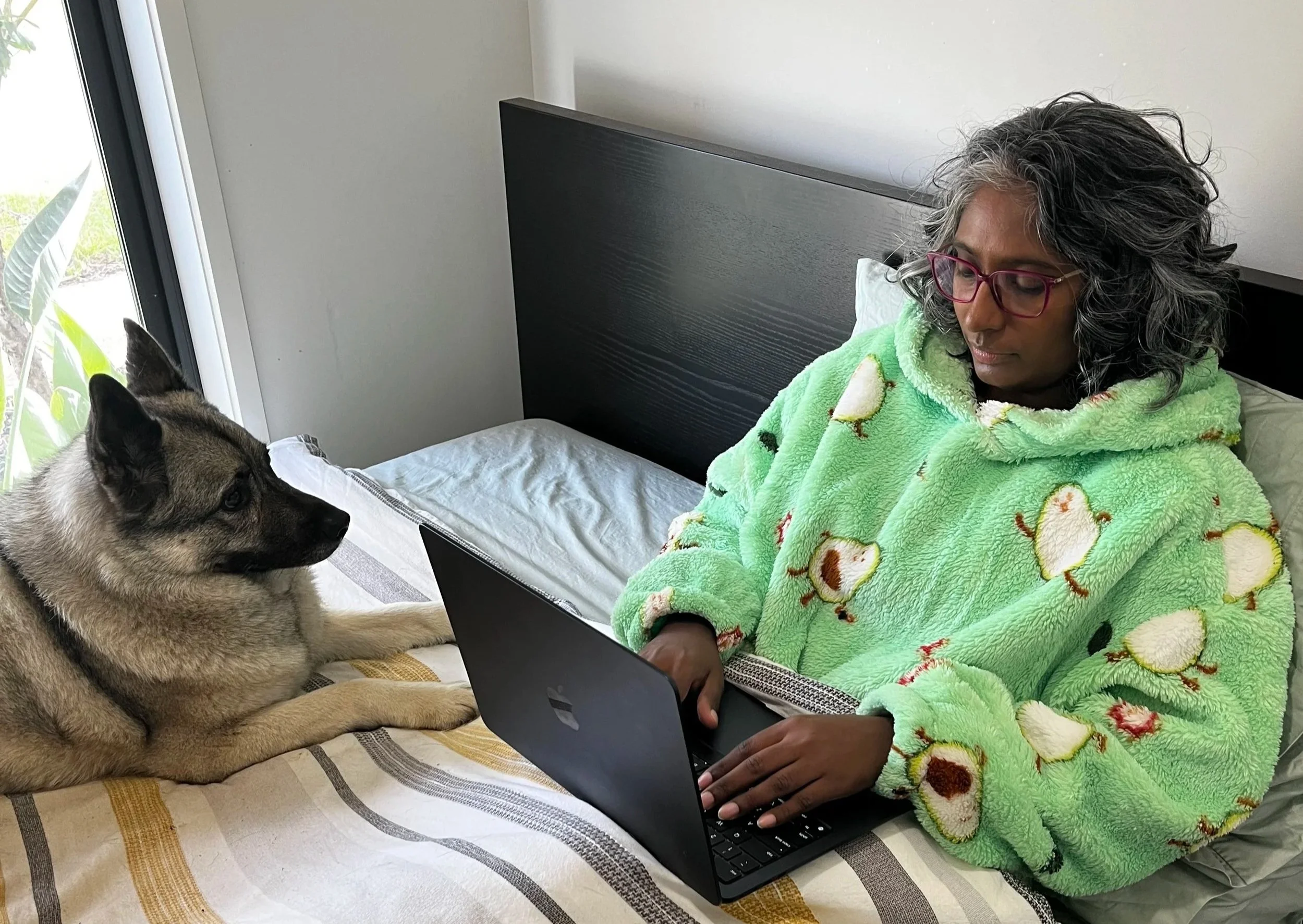 A woman with curly gray hair and glasses is sitting on a bed, using a laptop. She is wearing a green fleece blanket with a duck pattern. A cat with a black and gray coat is lying beside her, facing the laptop.