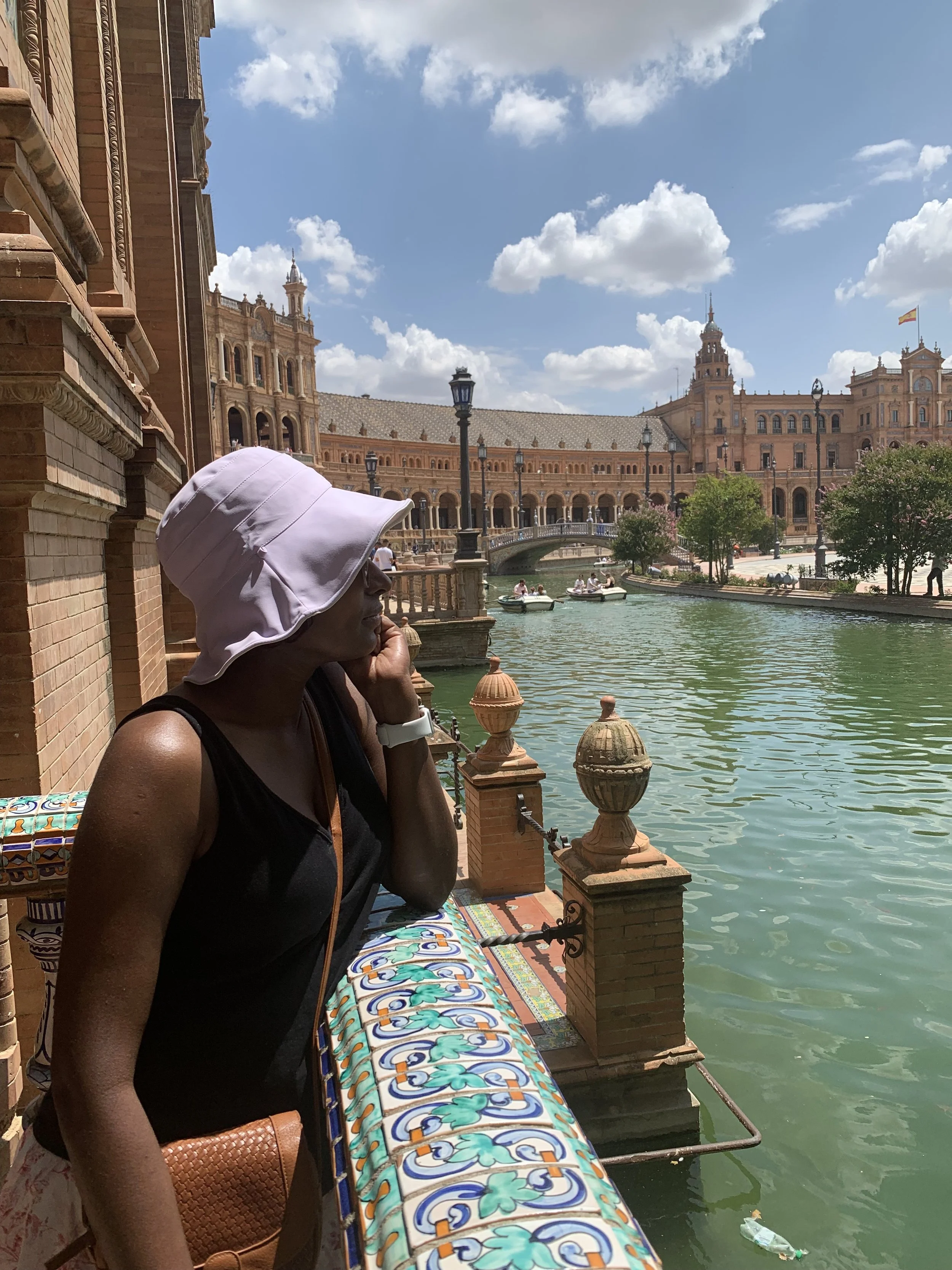 A woman with dark skin wearing a white sun hat and black sleeveless top, resting her chin on her hand while sitting by a decorative tiled railing overlooking a canal with small boats, historic buildings, and a bridge, under a partly cloudy sky.