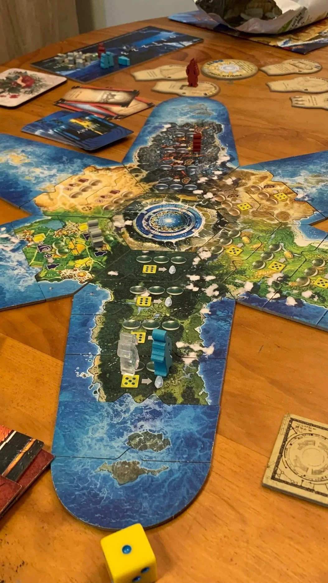 A tabletop game setup featuring a long game board with different themed sections, various game pieces including tokens, cards, dice, and miniatures, indicating an ongoing game session.