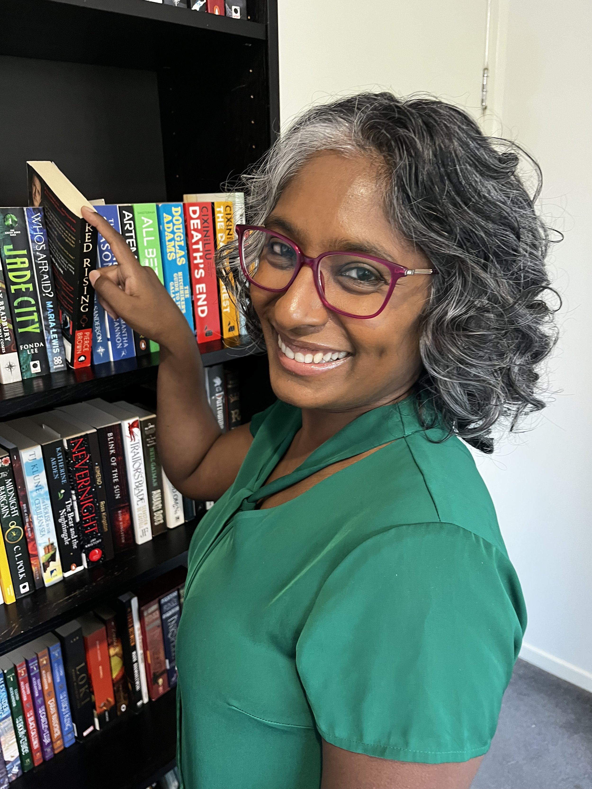 A woman with curly gray and black hair wearing pink glasses and a green shirt, smiling and pointing to a row of books on a black bookshelf.