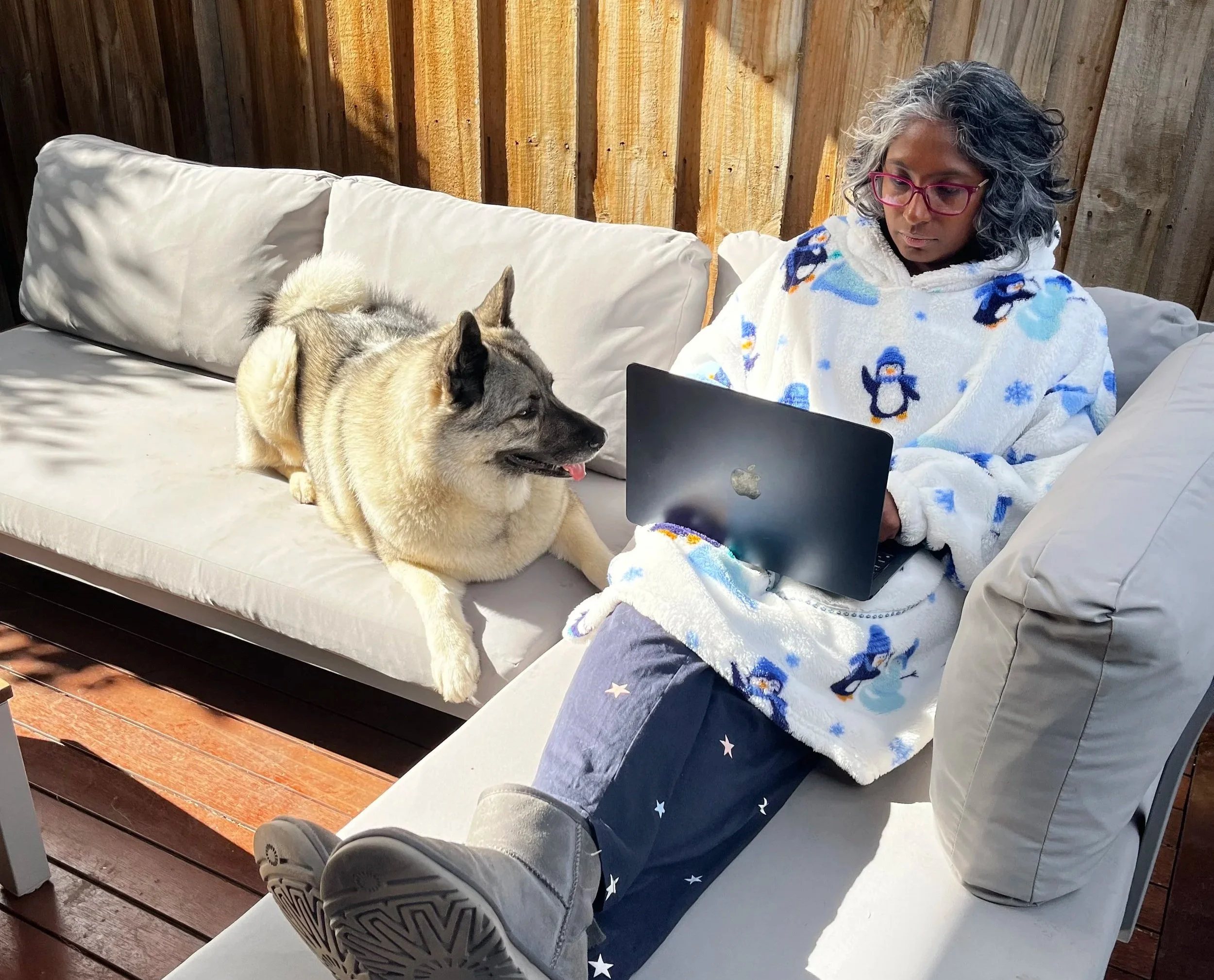 A woman with glasses and gray curly hair sitting on a sofa, working on a laptop, with a husky dog lying next to her. She is wearing a white fleece jacket with penguins and snowflakes. The setting appears to be a cozy indoor or covered outdoor space with a wooden wall and deck.