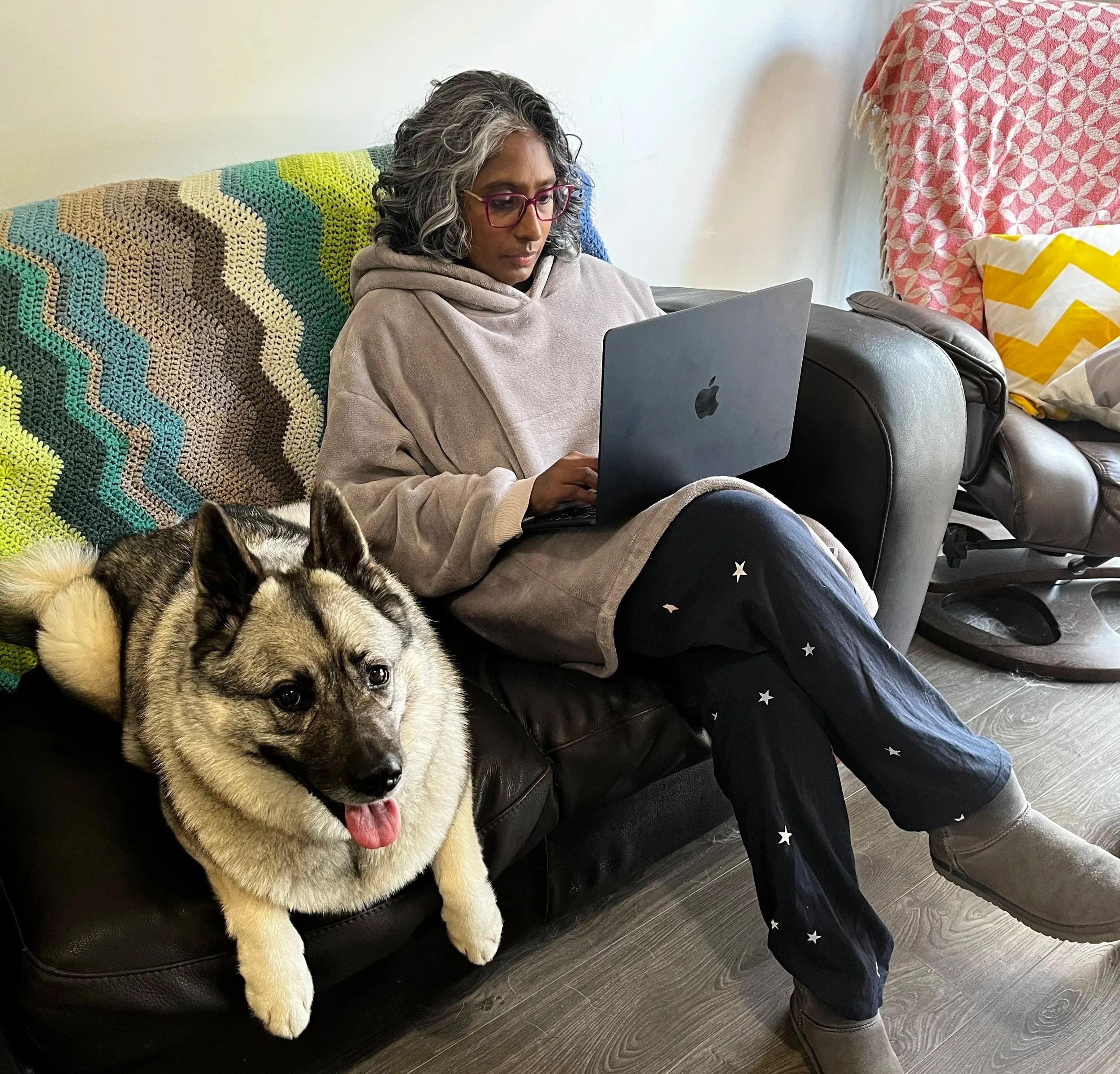 A woman with gray curly hair and glasses sitting on a couch with a colorful crocheted blanket, working on a laptop, and a puppy with a gray and white coat lying in front of her.