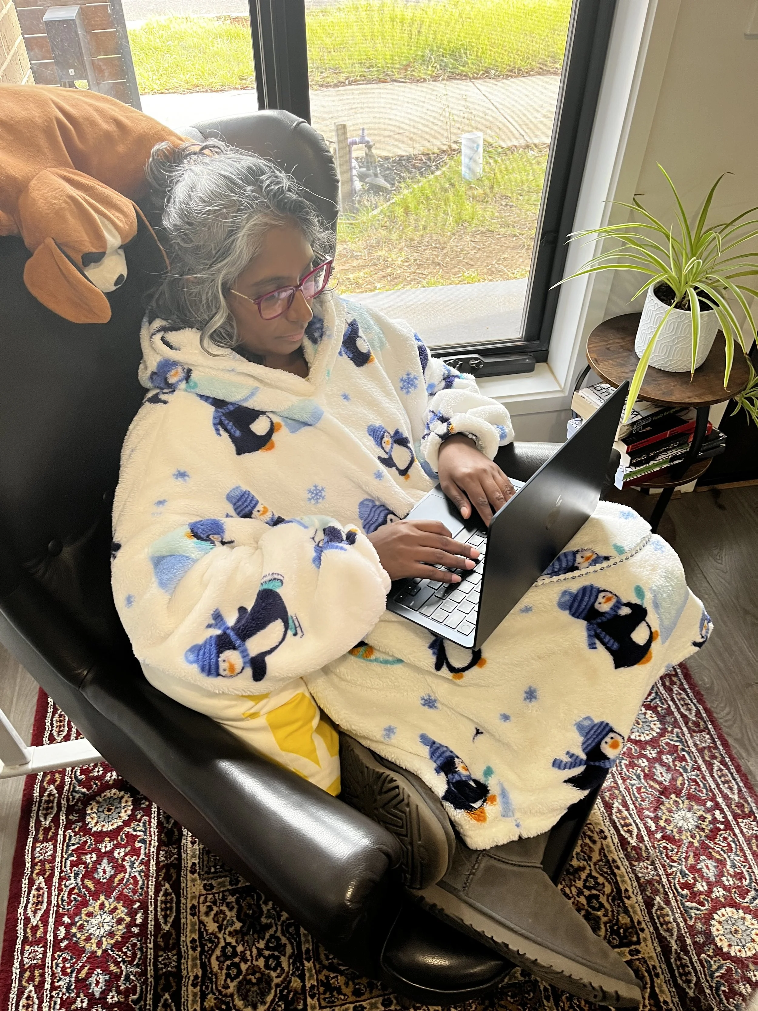 A woman with gray hair, glasses, and a penguin-patterned fleece blanket sitting on a black recliner, working on a laptop, near a large window with a view of grass and concrete outside, on a patterned rug with a side table holding a potted plant and books.