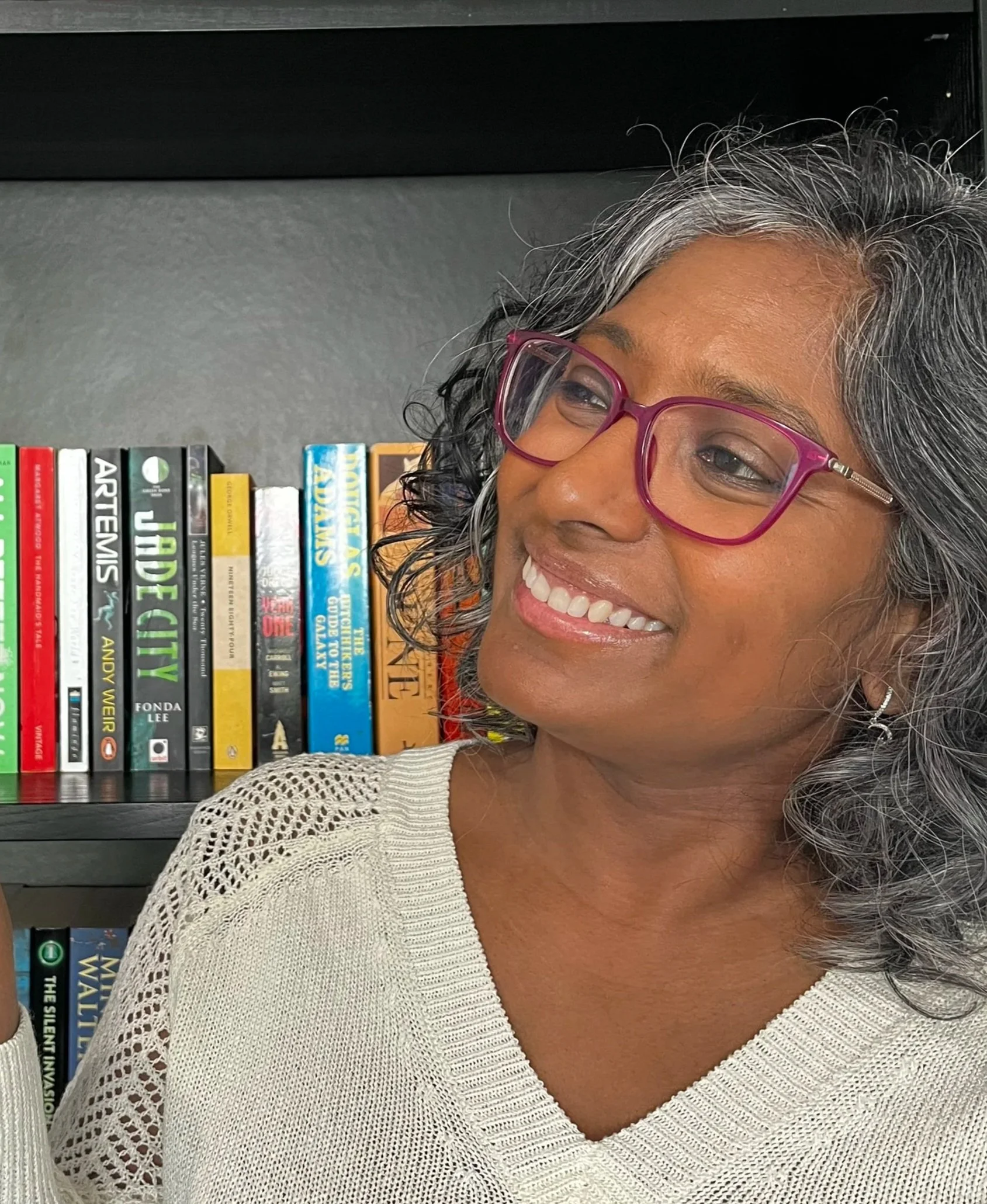A woman with gray and black curly hair, wearing pink glasses, smiling, in front of a bookshelf filled with colorful books.