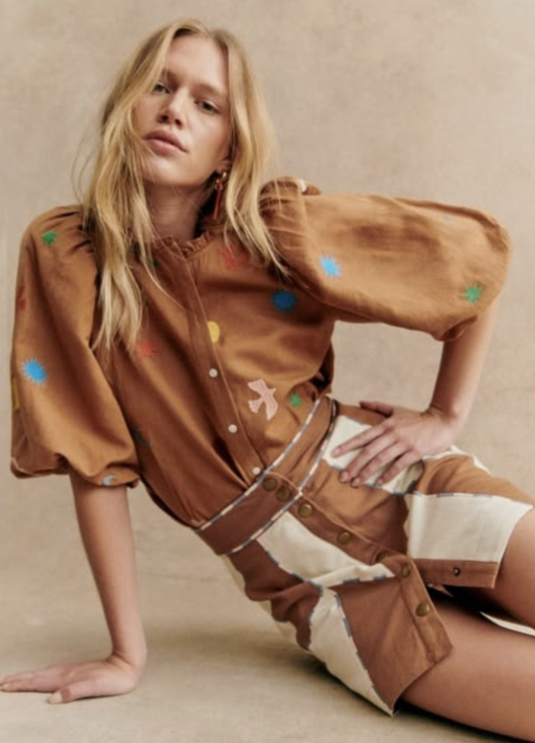 A young woman with long blond hair sitting on the ground with one hand on the floor and the other on her hip. She is wearing a brown blouse and shorts with colorful patches and has a confident expression.