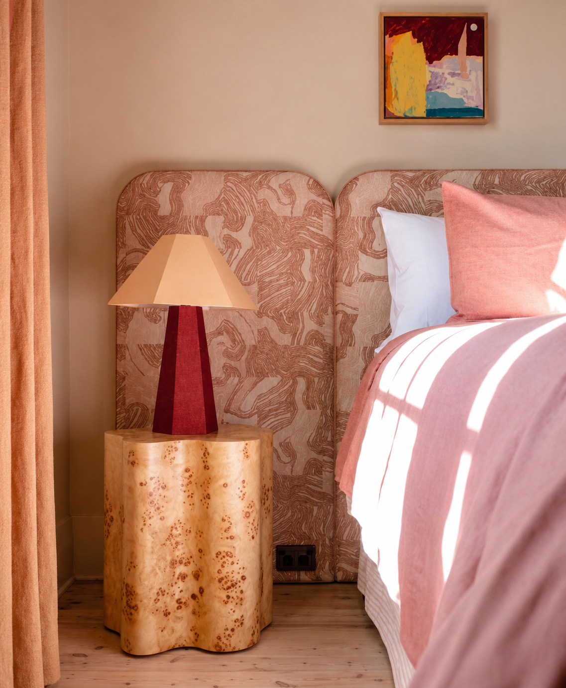 A cozy bedroom corner featuring a bed with pink bedding, a matching pink pillow, a bedside table with a red and beige lamp, patterned headboard, and a colorful abstract painting above the bed.