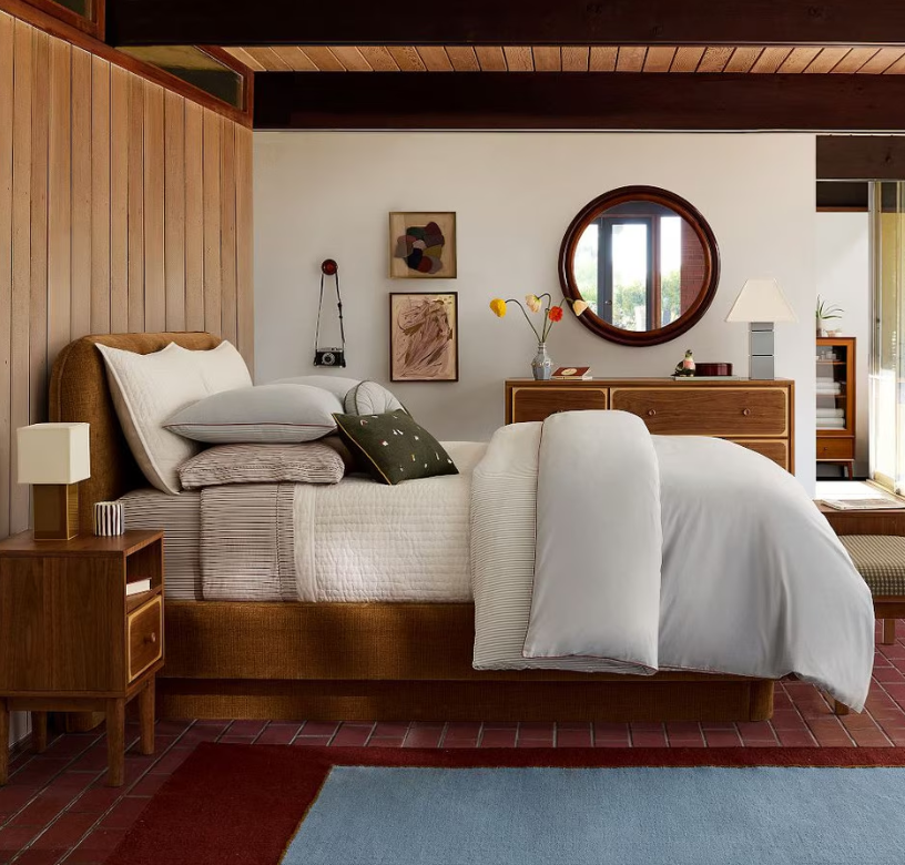 A cozy bedroom with a large bed featuring white linens and decorative pillows, wooden furniture including a nightstand with a lamp, a dresser with a flower vase and a mirror above, and wall art, with natural light coming through a window or door.