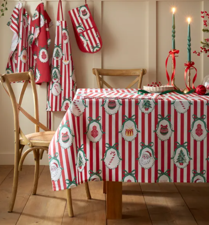 Dining table covered with a red and white striped Christmas-themed tablecloth featuring ornaments, Santa, and Christmas trees. Hanging on the wall are matching holiday towels. Two lit green candles with red ribbons are on the table. Wooden chairs surround the table, and the floor is wooden.