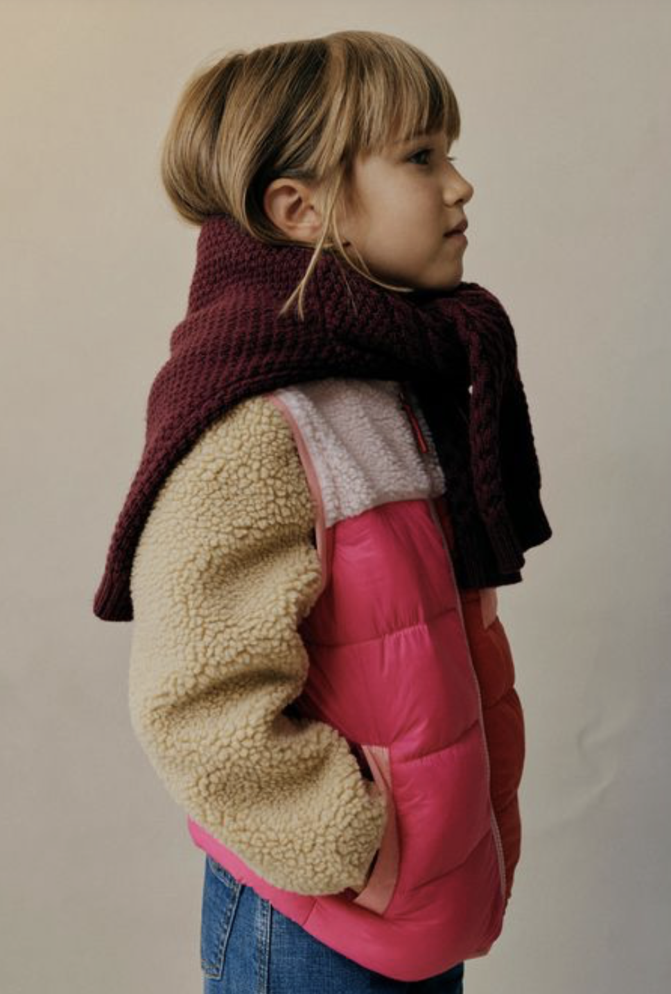Profile of a young girl with blonde hair wearing a pink puffer jacket, a cream-colored fleece sweatshirt, and a maroon woven scarf, standing against a plain beige wall.