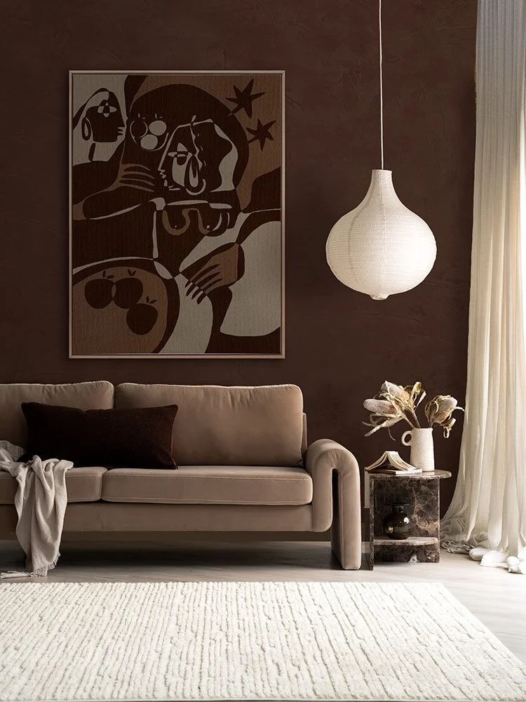 Living room with beige sofa, black pillow, beige throw blanket, abstract art on dark brown wall, hanging white light fixture, side table with vase and flowers, cream curtains, light-colored rug.