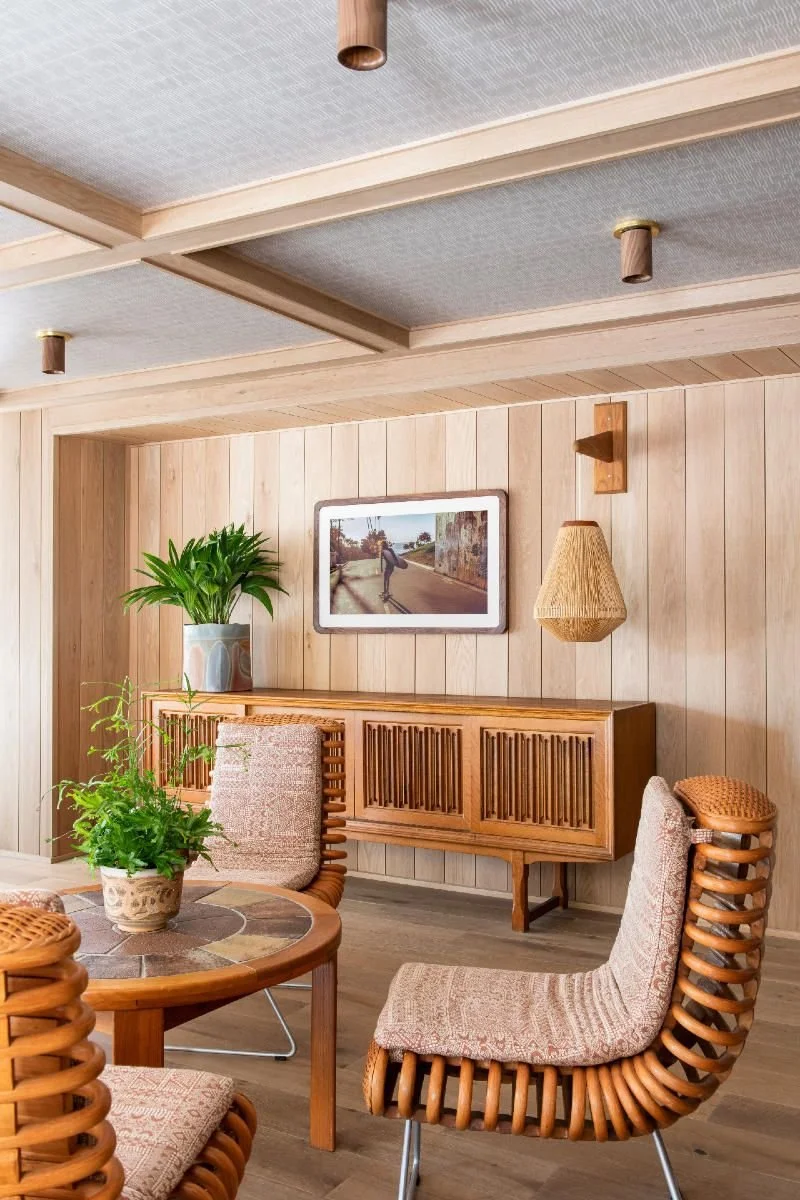 A cozy living room corner with wooden furniture, including a round table with a potted plant, cushioned chairs with rattan frames, a wooden sideboard, and wall decor with a photograph and a wicker lamp, all featuring natural wood paneling walls.