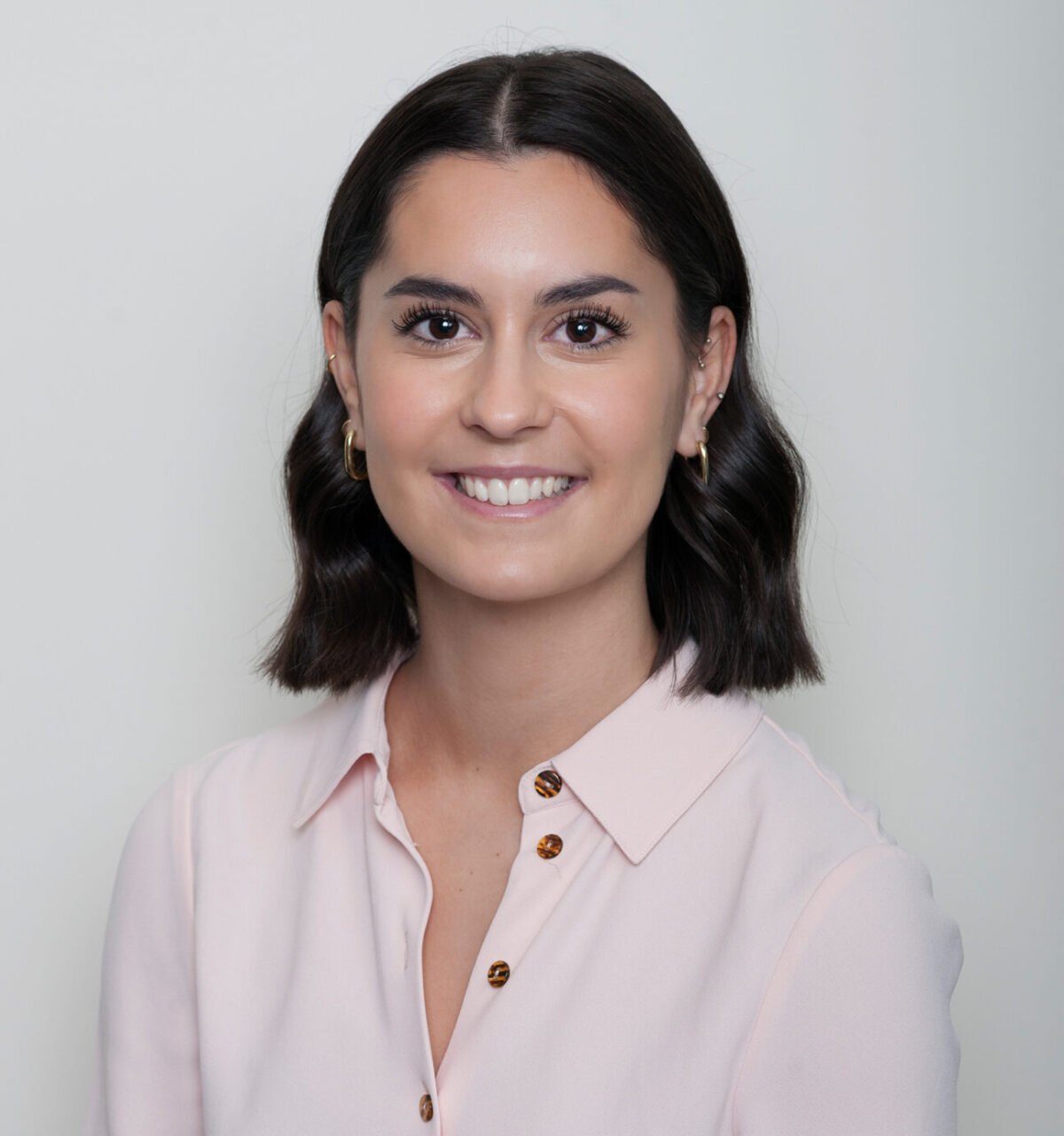 A young woman with shoulder-length dark hair, smiling at the camera, wearing a light pink blouse with button details, gold hoop earrings, and a light makeup look. The background is plain and neutral.