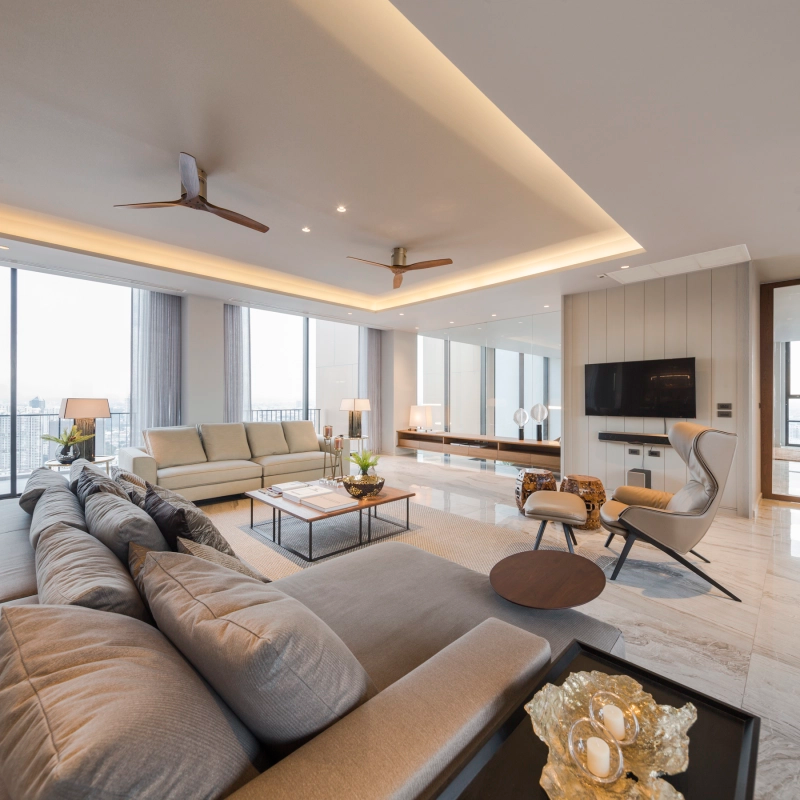 Bright modern living room with large windows, beige couches, a TV on the wall, a lounge chair, and soft lighting.