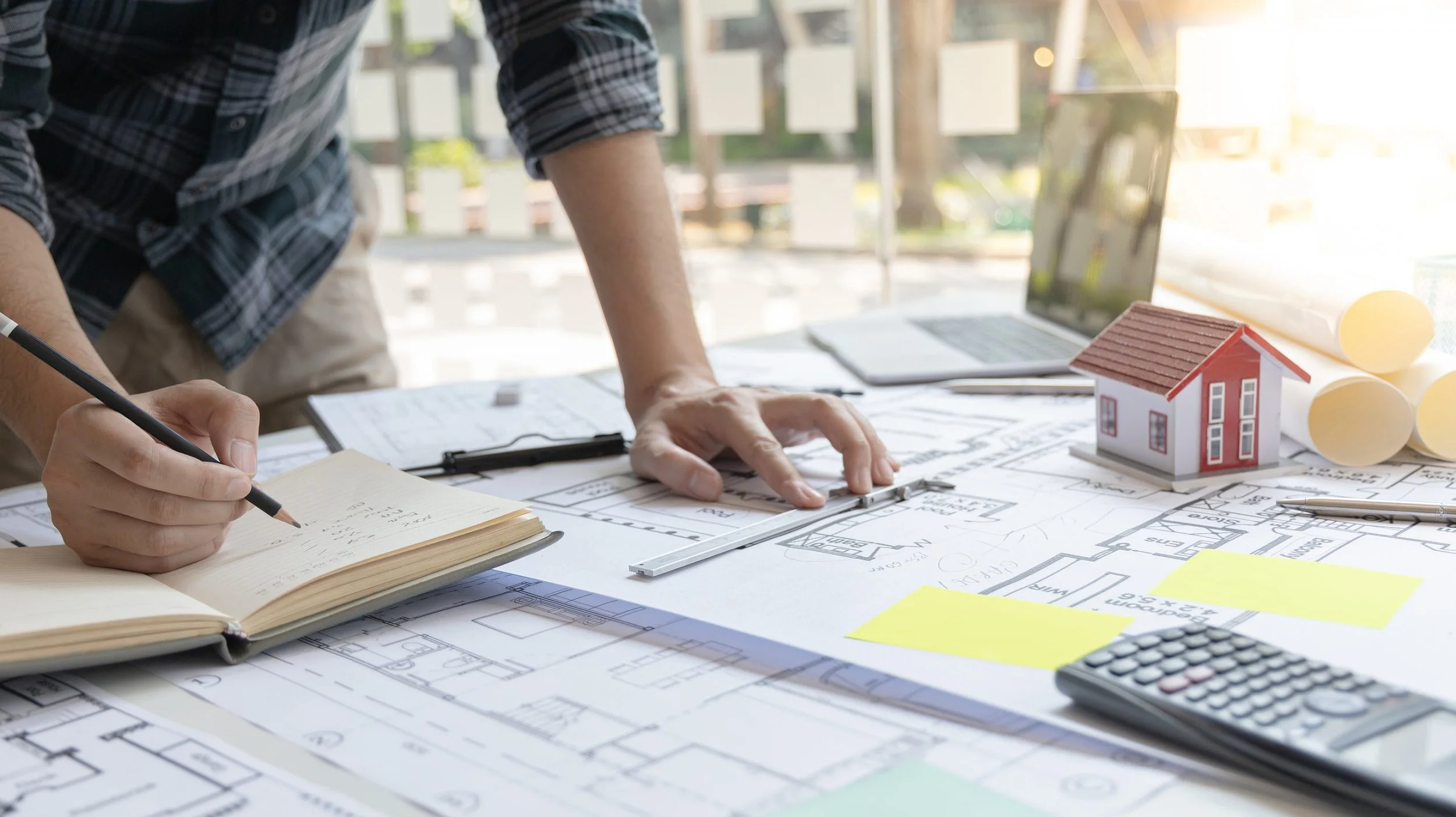 An architect working on building plans at a desk with a small house model, rolled-up blueprints, a notebook, a calculator, and a laptop in the background.