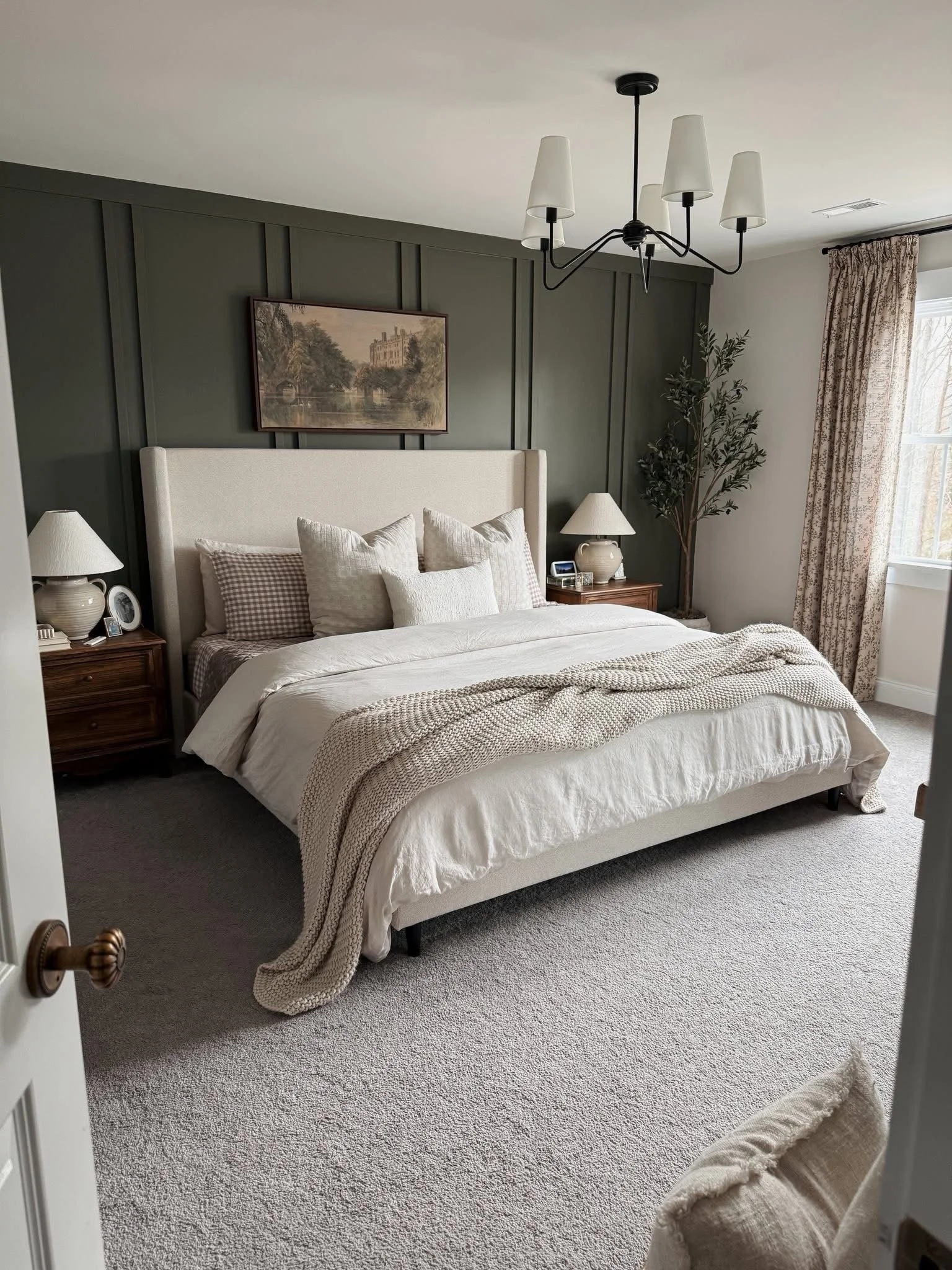 A cozy bedroom with a white upholstered bed, multiple pillows, and a beige knit throw blanket. There are two matching nightstands with lamps, a framed painting of a building across a river, a potted tree near a window with patterned curtains, and a black chandelier hanging from the ceiling.