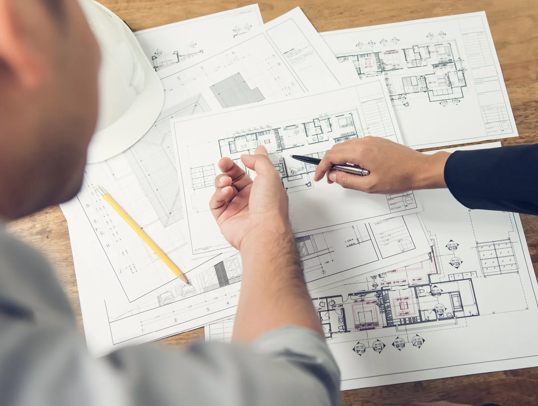 Two people reviewing detailed architectural blueprints laid out on a wooden table, one pointing at a specific section with a pen, and a hard hat and pencil also on the table.