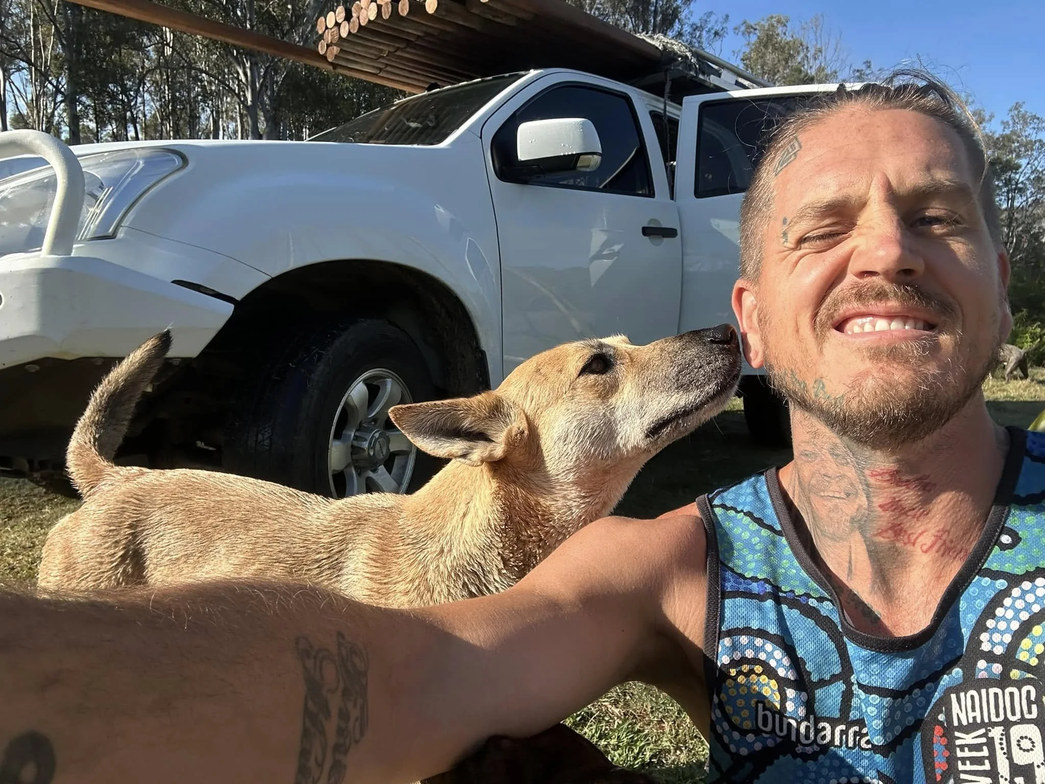 On country a good looking, wise man sits, smiling while taking a selfie with a dingo dog and a white ute work truck in the background, outdoors on a sunny day.