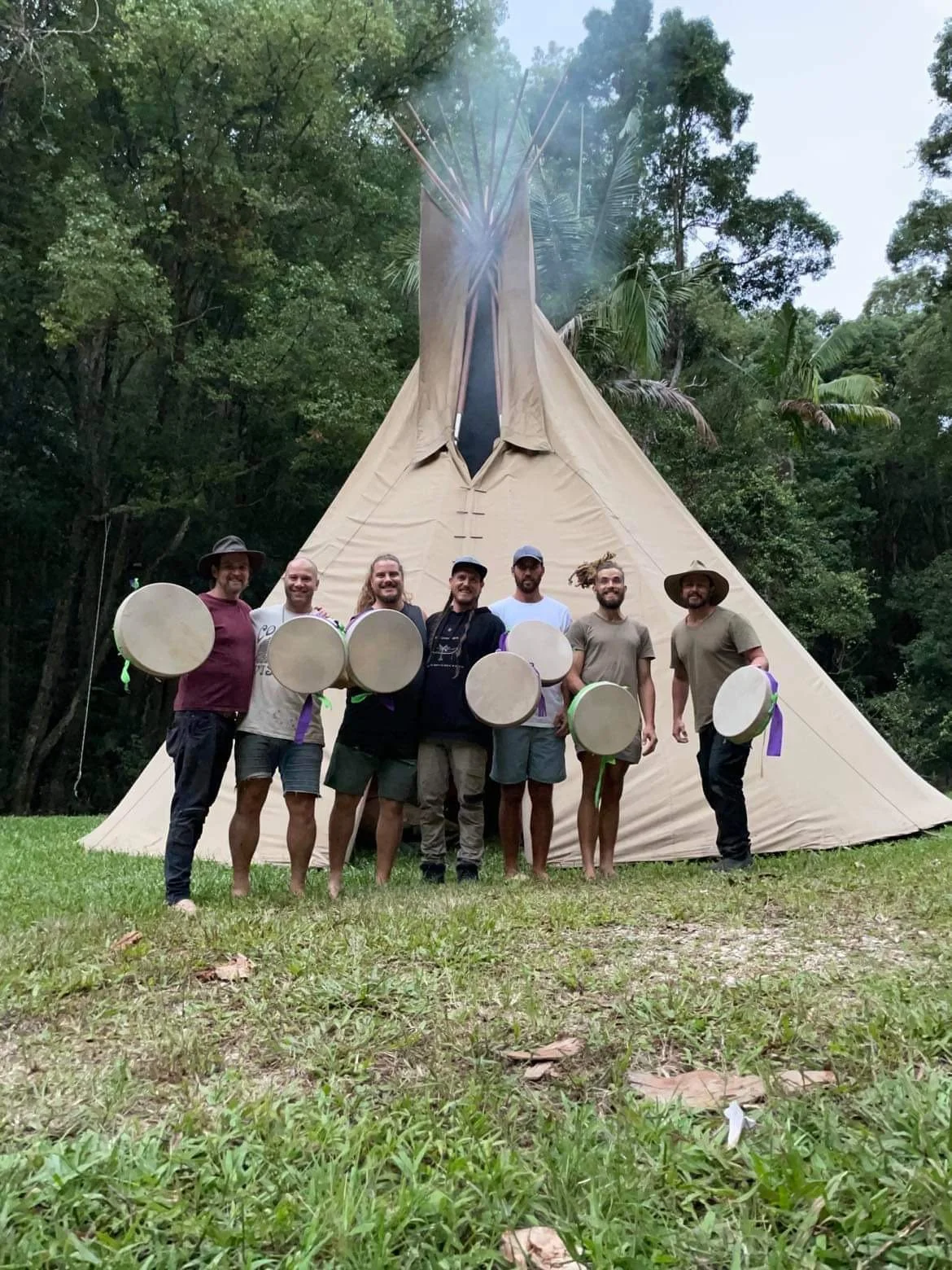 Group of men standing in front of a tipi teepee, holding round frame drums, shamanic drums after a drum making workshop, ceremony, rite of passage and cultural programs sharing traditional knowledge at an outdoor event.