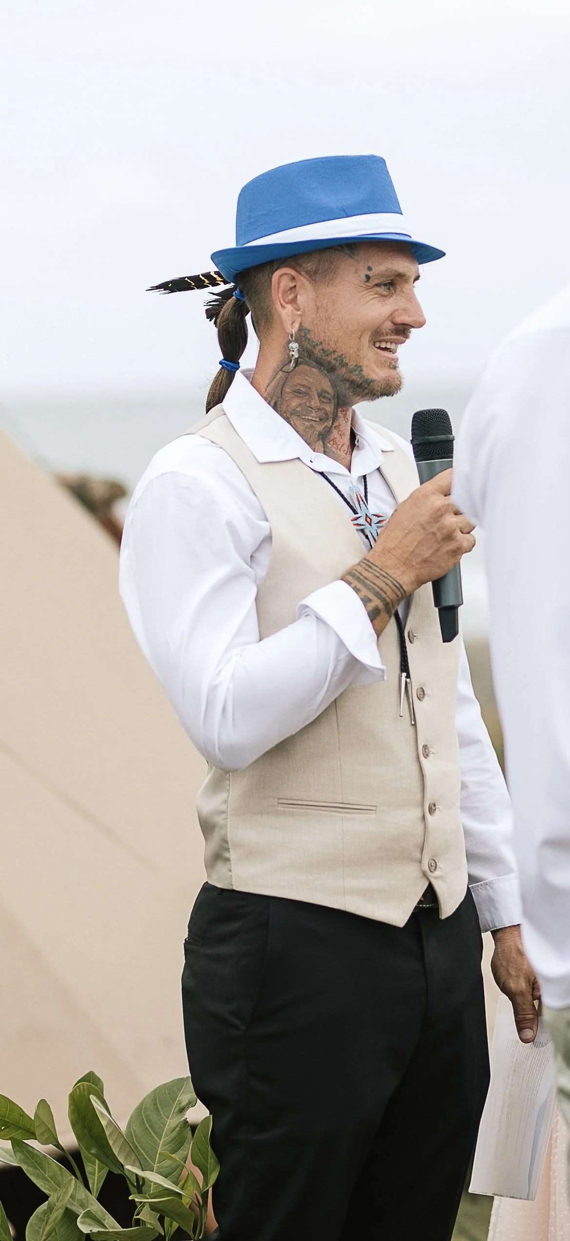 Good looking well dressed man with tattoos and piercings wearing a feathered shamanic hat speaks wisdom, shares knowledge and mentors through ceremony, workshops, cultural programs and rites of passage at an outdoor event. Sobriety, and spiritual