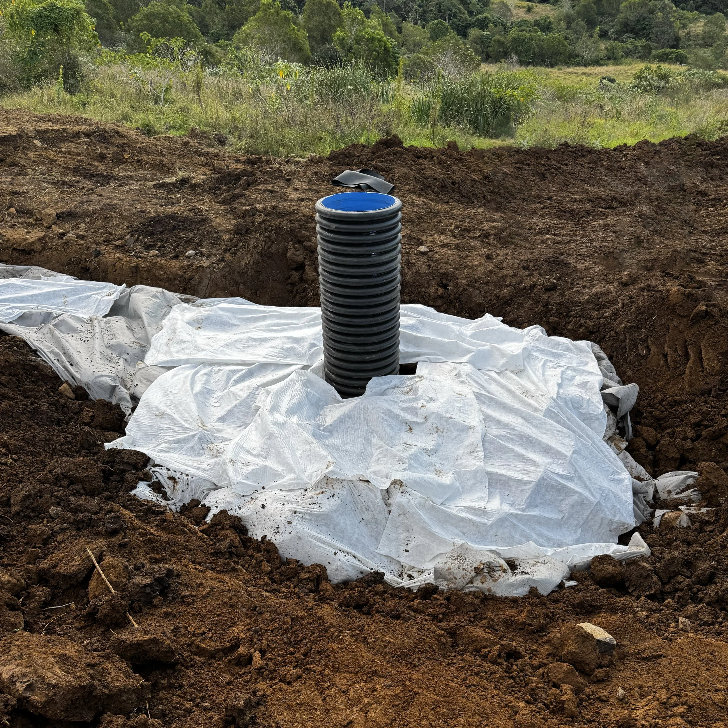 Black corrugated drainage pipe with a blue opening installed in freshly excavated soil, surrounded by white geotextile fabric and green vegetation.
