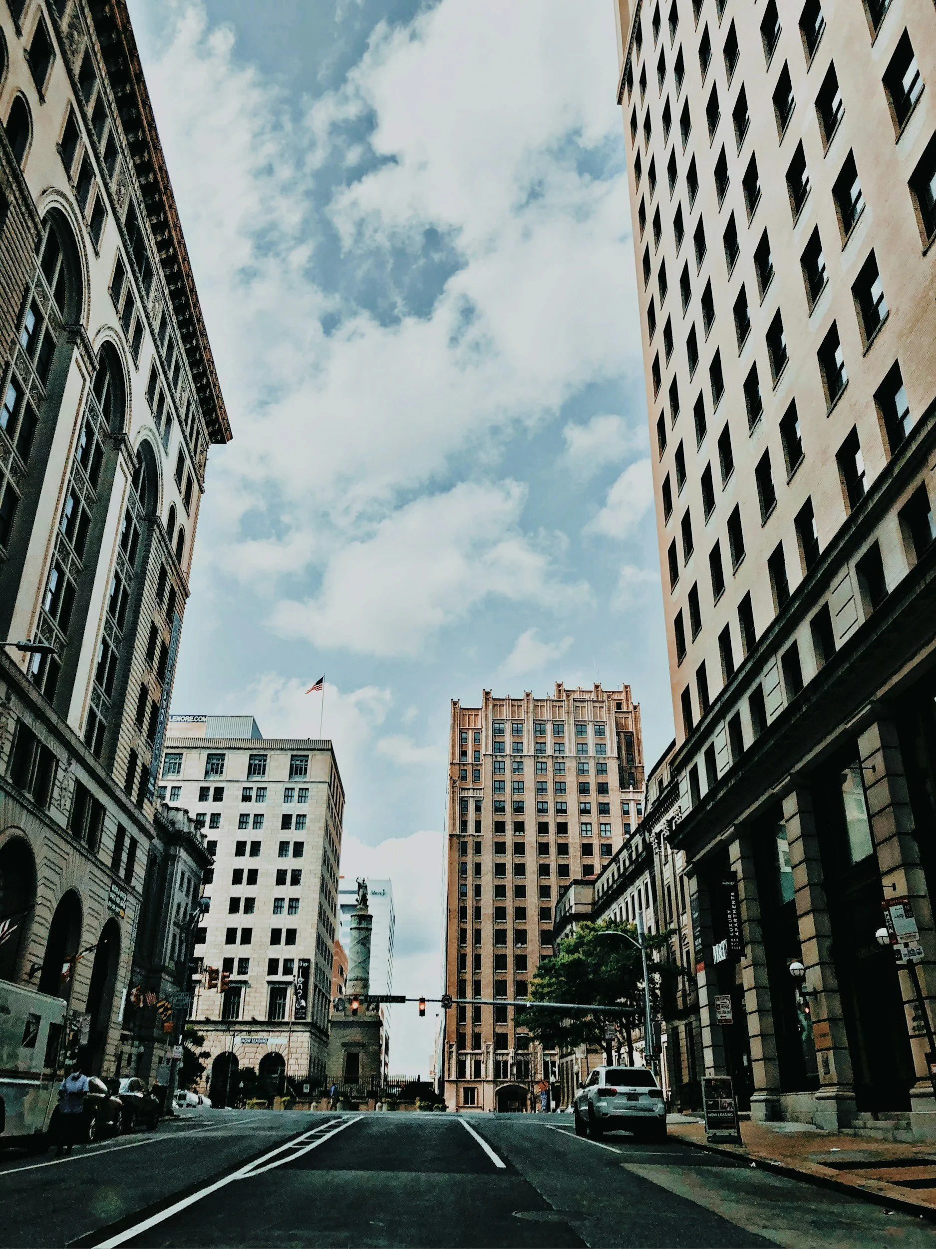 A city street scene with tall buildings on both sides, cars parked along the curb, and an American flag on a pole in the background. Traffic lights are visible at the intersection, and the sky is partly cloudy.