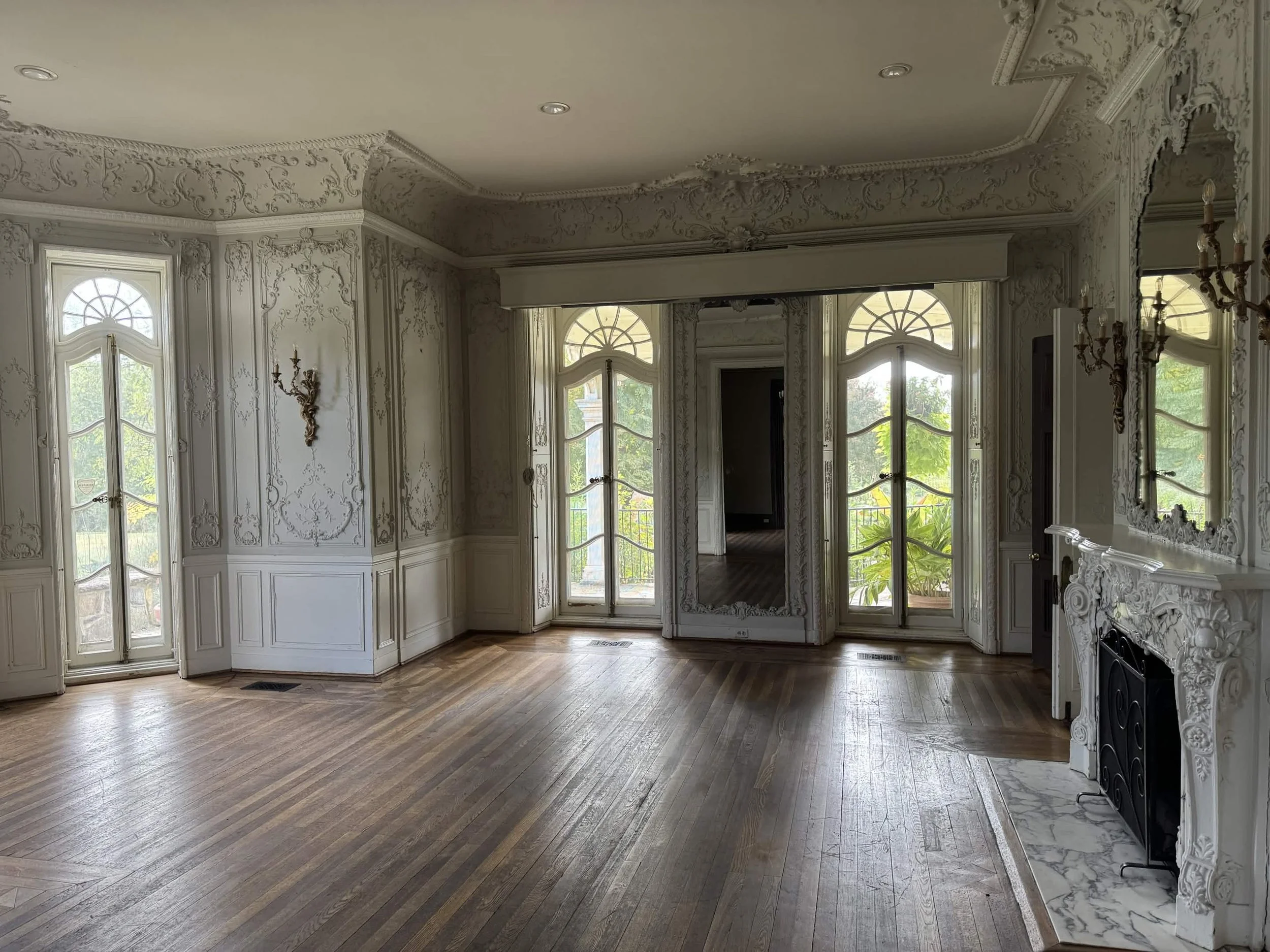 An elegant room with ornate white crown molding, decorative wall paneling, and a fireplace. Large windows with curved tops let in natural light, and the room has hardwood floors.