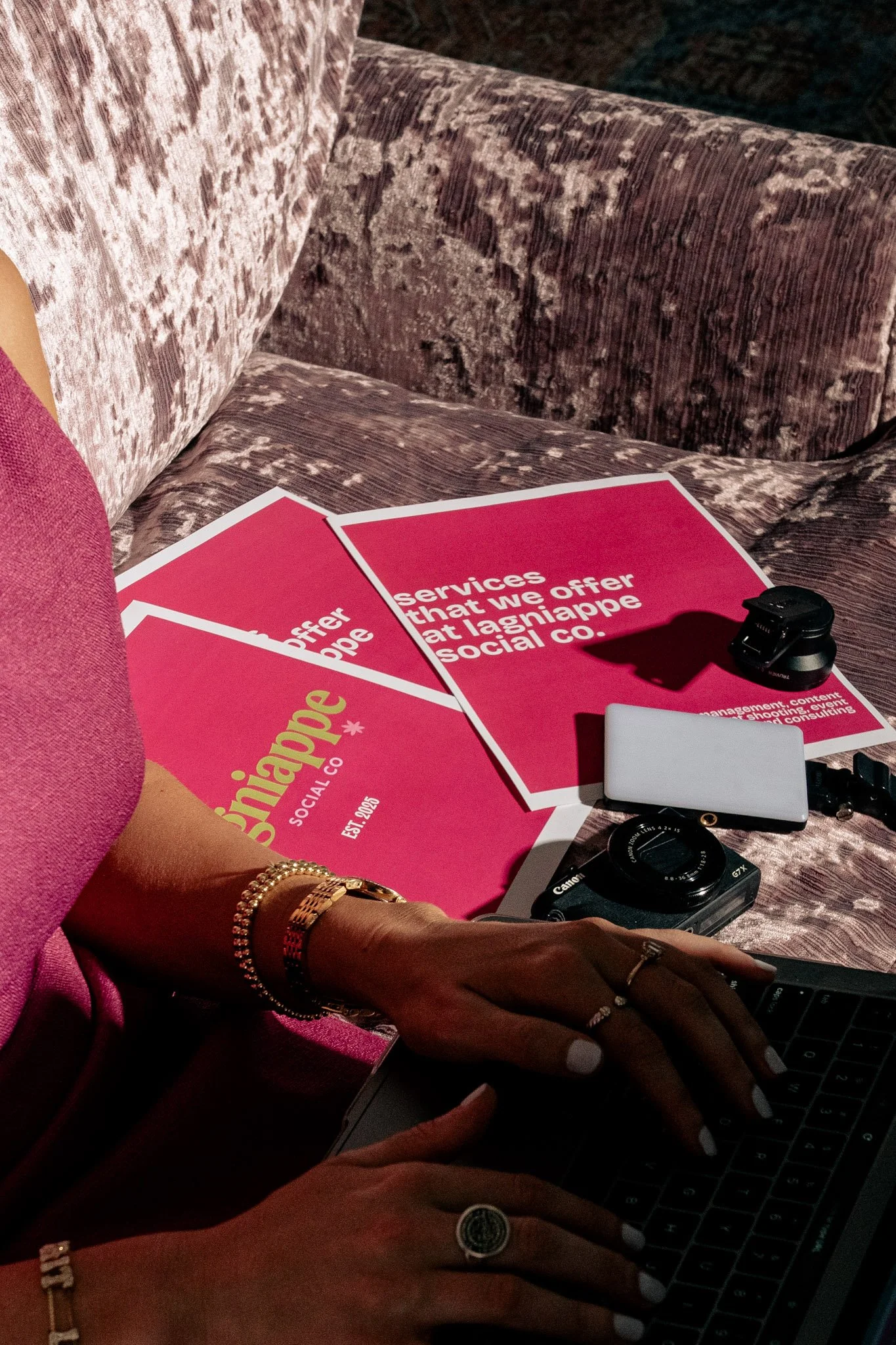 Close-up of a person's hands typing on a laptop keyboard, with orange and gold jewelry on their wrists and fingers. Several flyers with pink and white colors, displaying promotional or informational content, are on a patterned couch or table surface.