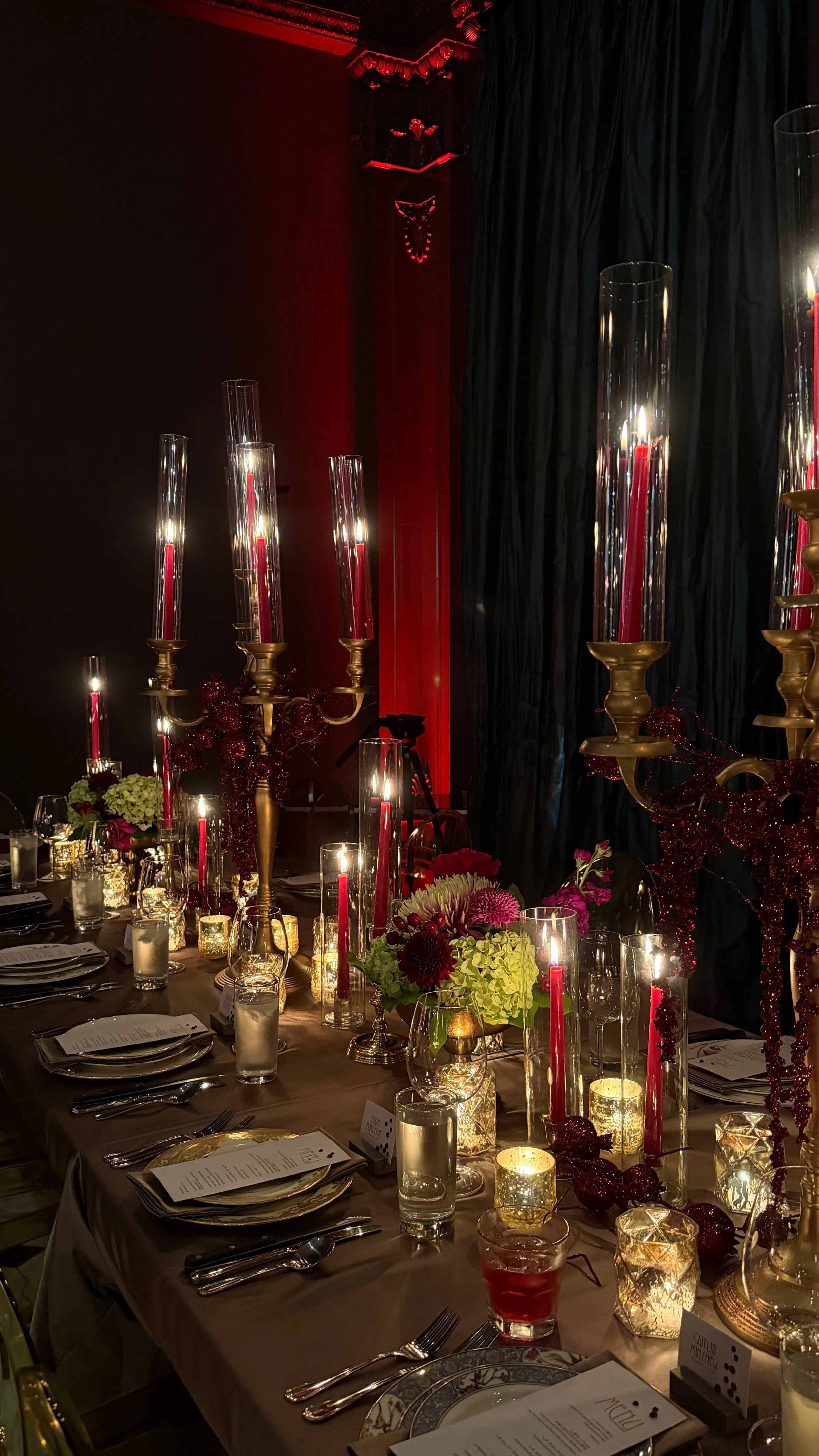 Elegant dinner table decorated with tall red candles in glass holders, floral arrangements, lit candles, and gold candelabras, set for a formal event or celebration in a dimly lit room with dark curtains and ornate trim.