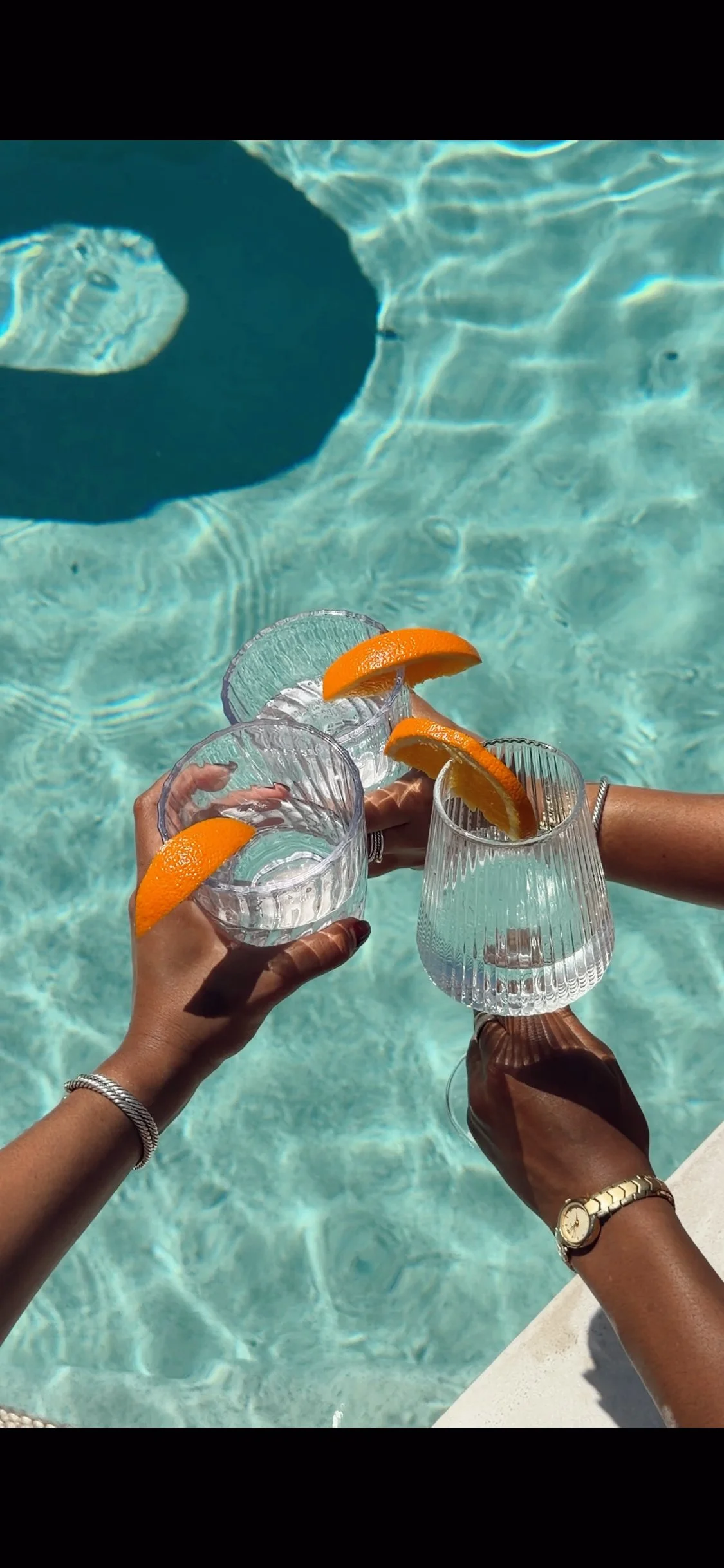 Three people holding glass glasses with orange slices by a swimming pool, with sunlight reflecting on the water.