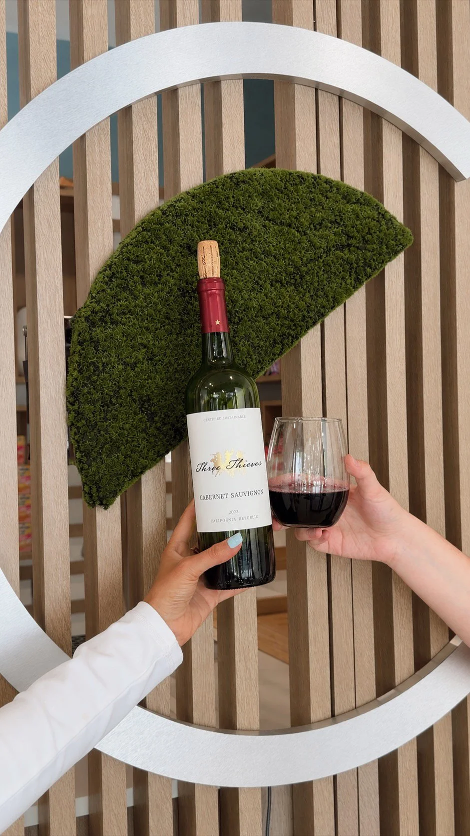 Two hands holding a wine glass and a bottle of red wine in front of a wooden decorative wall with semi-circular greenery and a white curved accent.