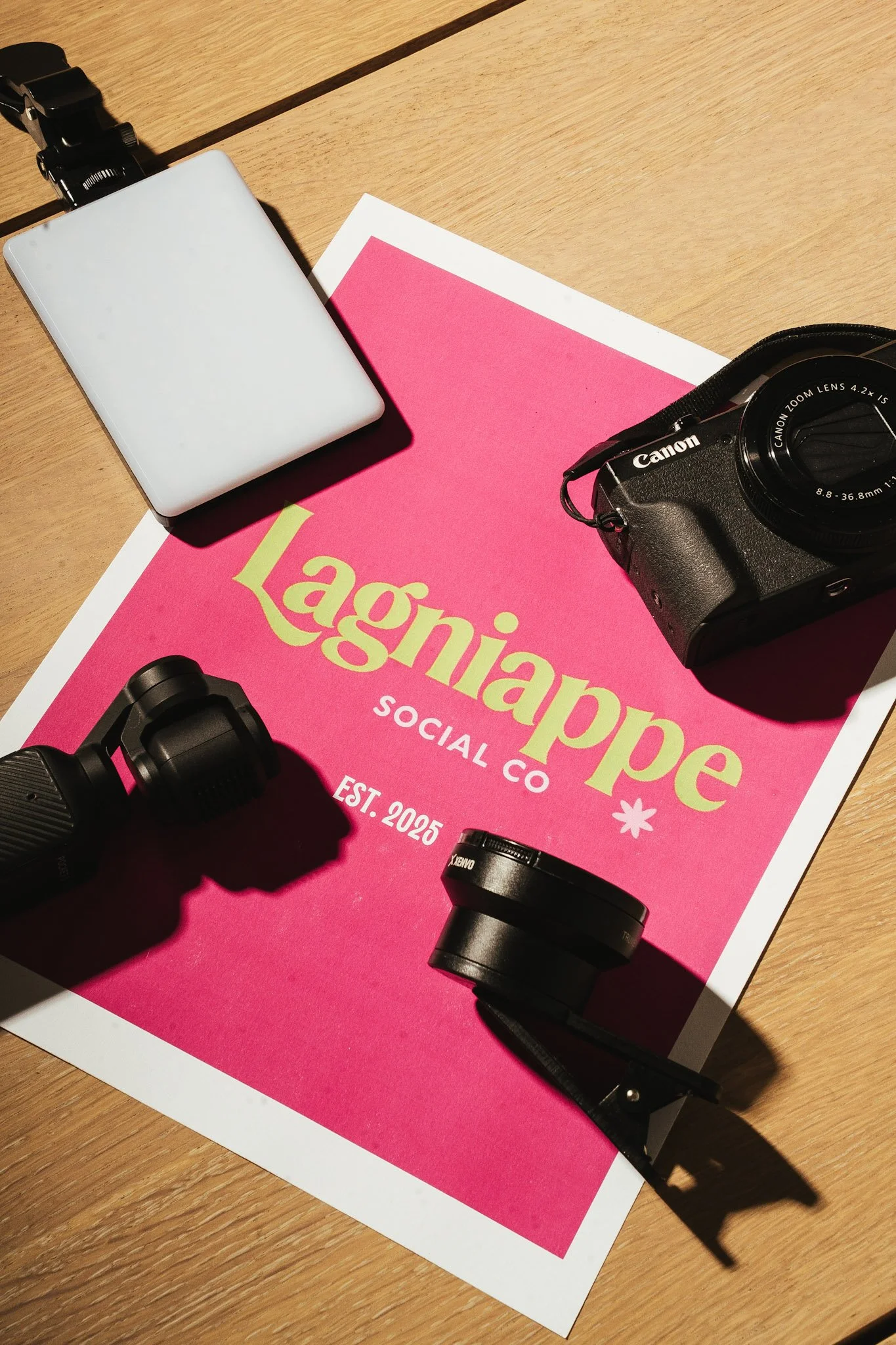 A desk with a pink and green magazine titled 'Lagliane' social co, a white external hard drive, a black DSLR camera, and camera lenses on a wooden surface.