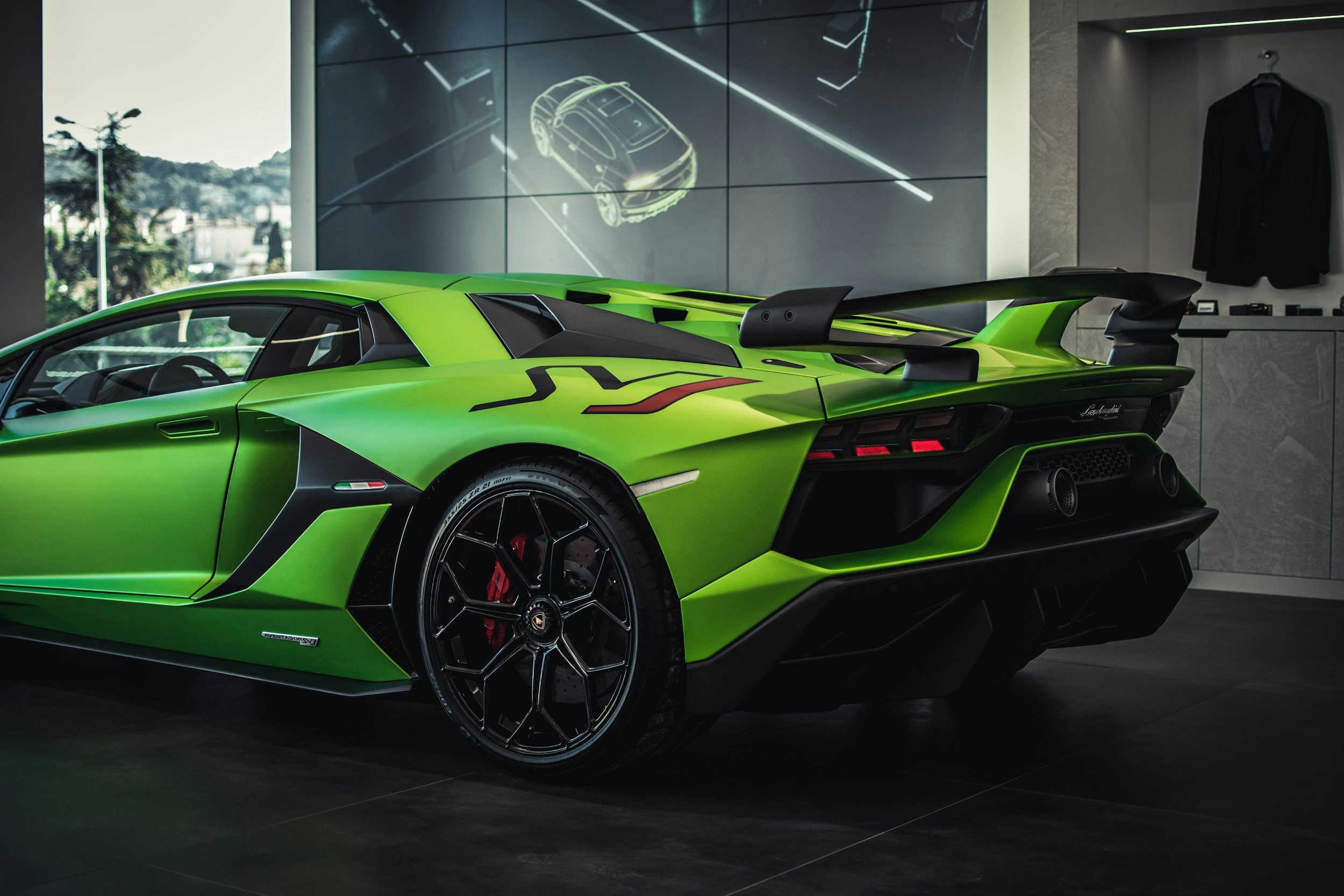 A bright green Lamborghini sports car in a showroom, with a large display screen behind it showing a digital outline of a car. The showroom has a modern design with gray walls, a black floor, and a suit hanging on the wall.