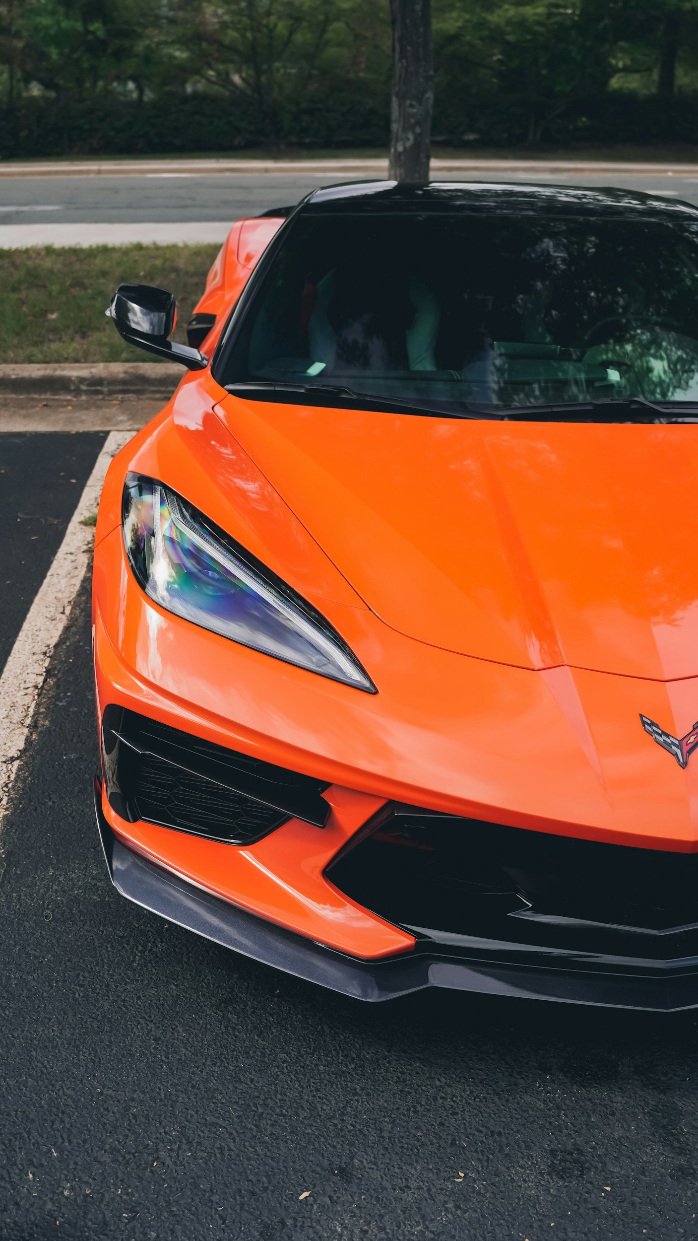 Close-up of an orange sports car parked in a parking lot, featuring a sleek front design with black accents and a detailed headlight.
