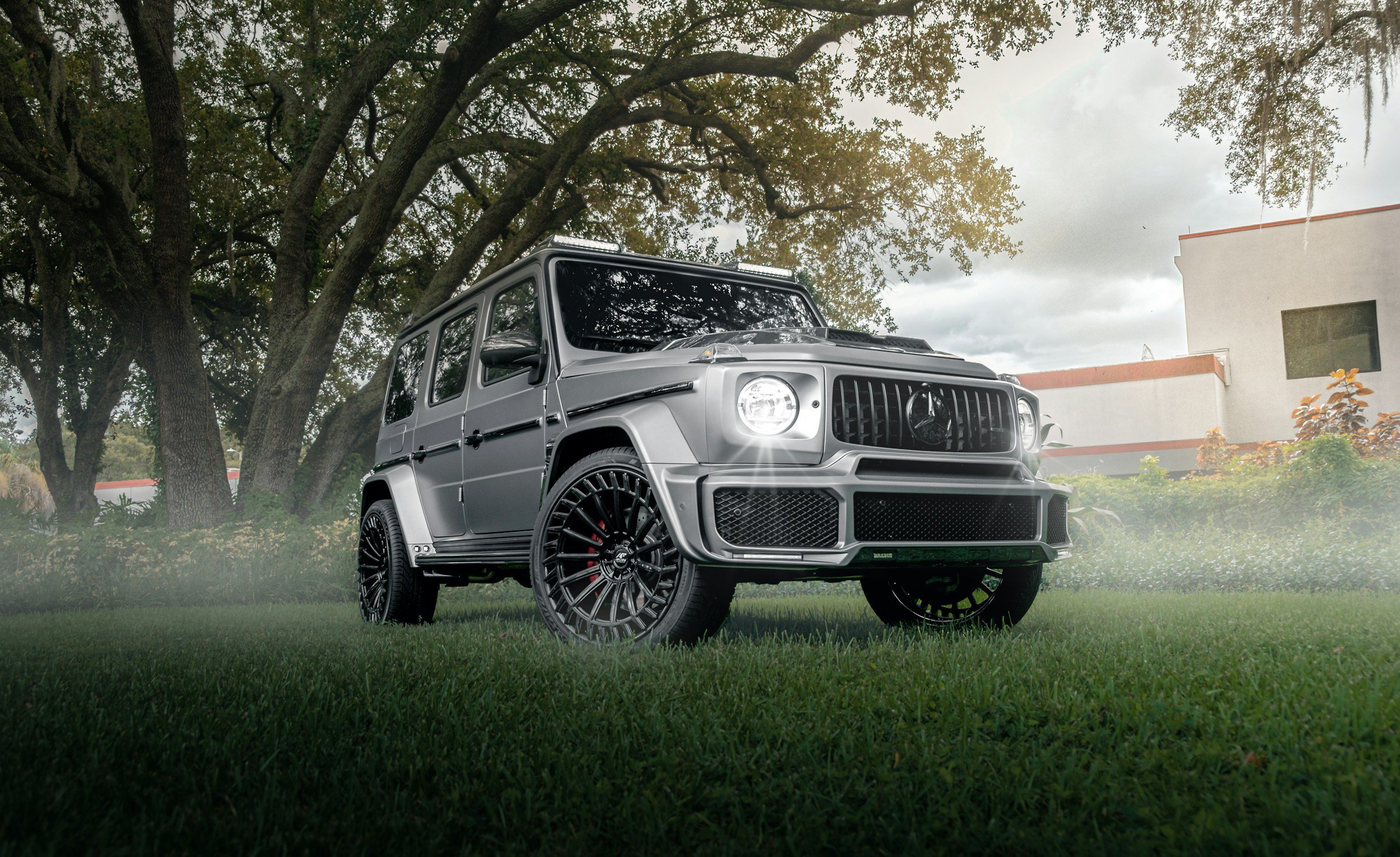 A silver Mercedes-Benz G-Class vehicle parked on a lawn under large trees with thick branches, with a building in the background. The vehicle has custom black rims and is positioned at an angle.