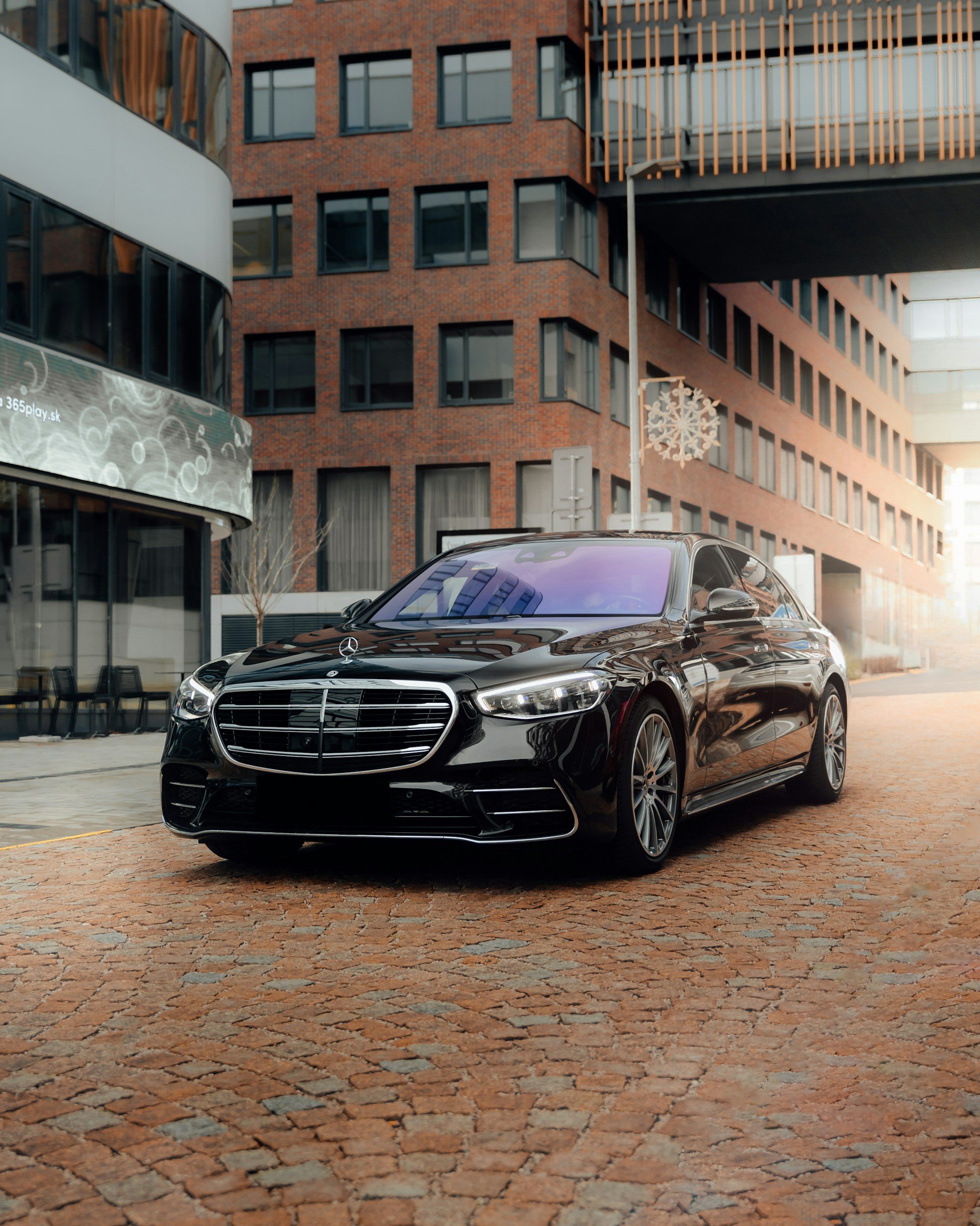 Black Mercedes-Benz car parked on cobblestone street in front of modern brick and glass building with hanging snowflake decoration