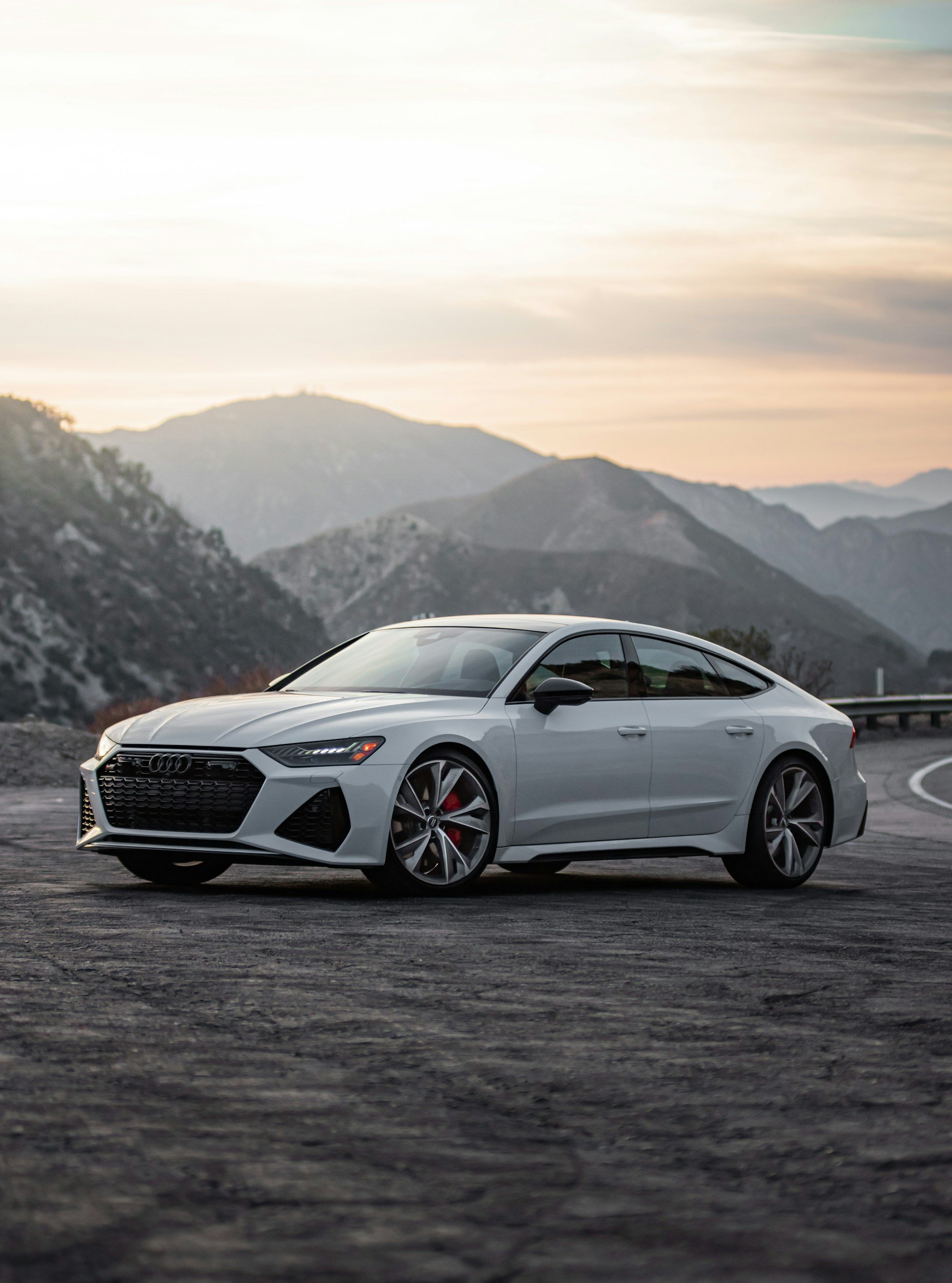 A silver sports car, likely an Audi, parked on a scenic overlook with mountains in the background and a sunset sky.