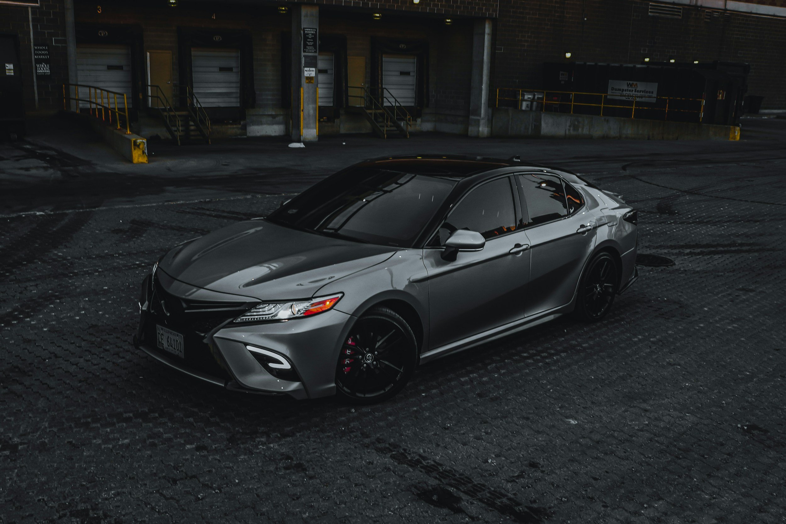 A silver sports car parked in an industrial parking lot at night, with a dark brick building and loading dock doors in the background.