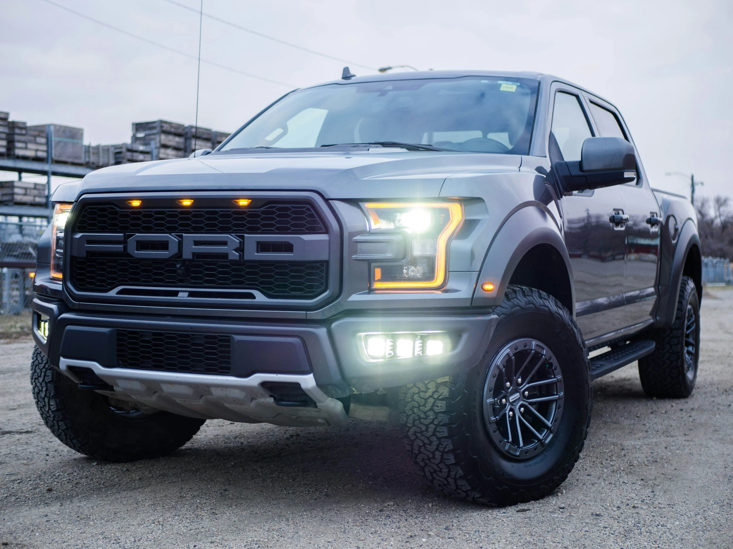 A silver Ford truck with bright headlights on, parked outdoors on a gravel surface, with blurry storage racks or pallets in the background.