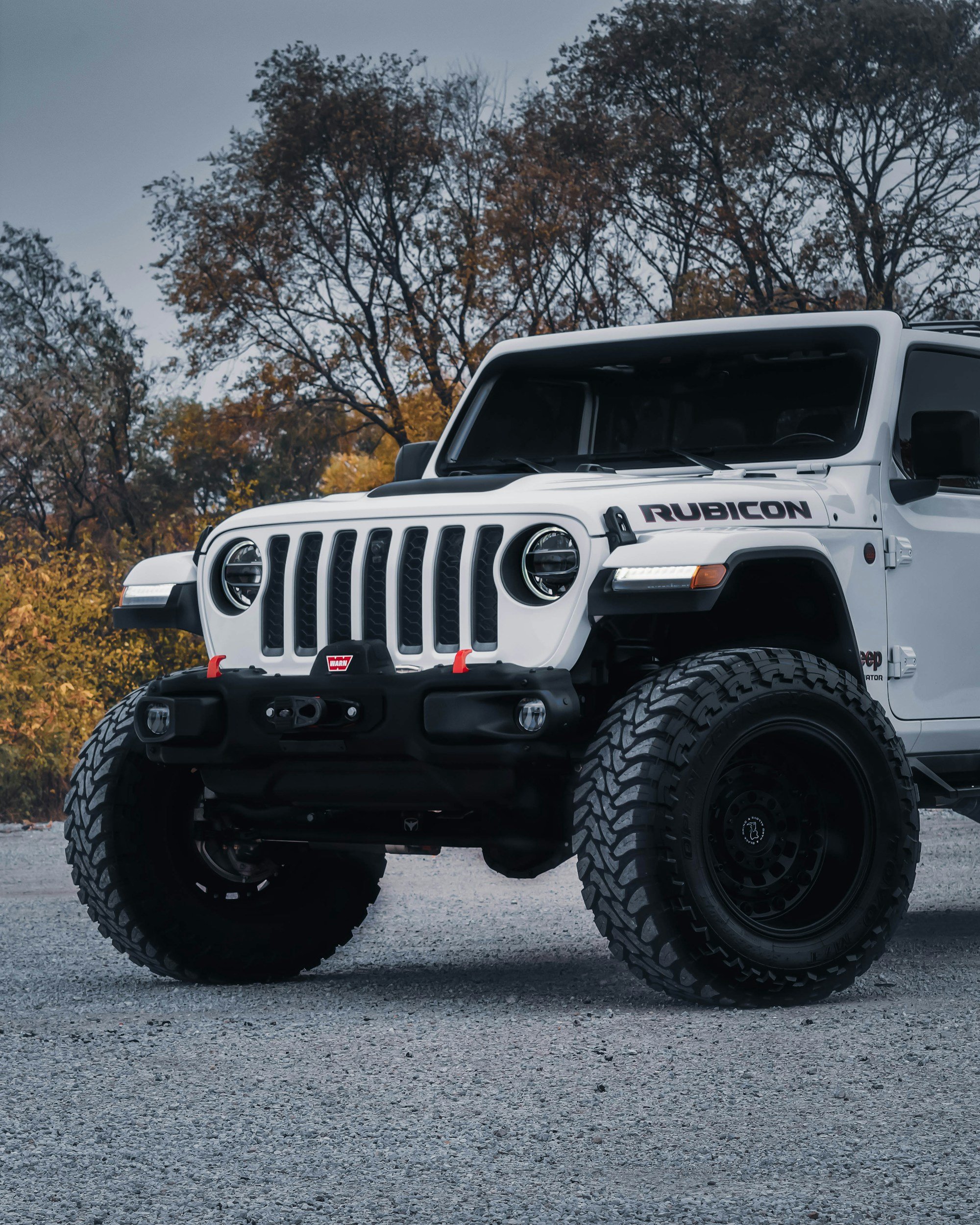 Close-up of a white Jeep Rubicon with large off-road tires parked on a gravel surface, autumn trees in the background.