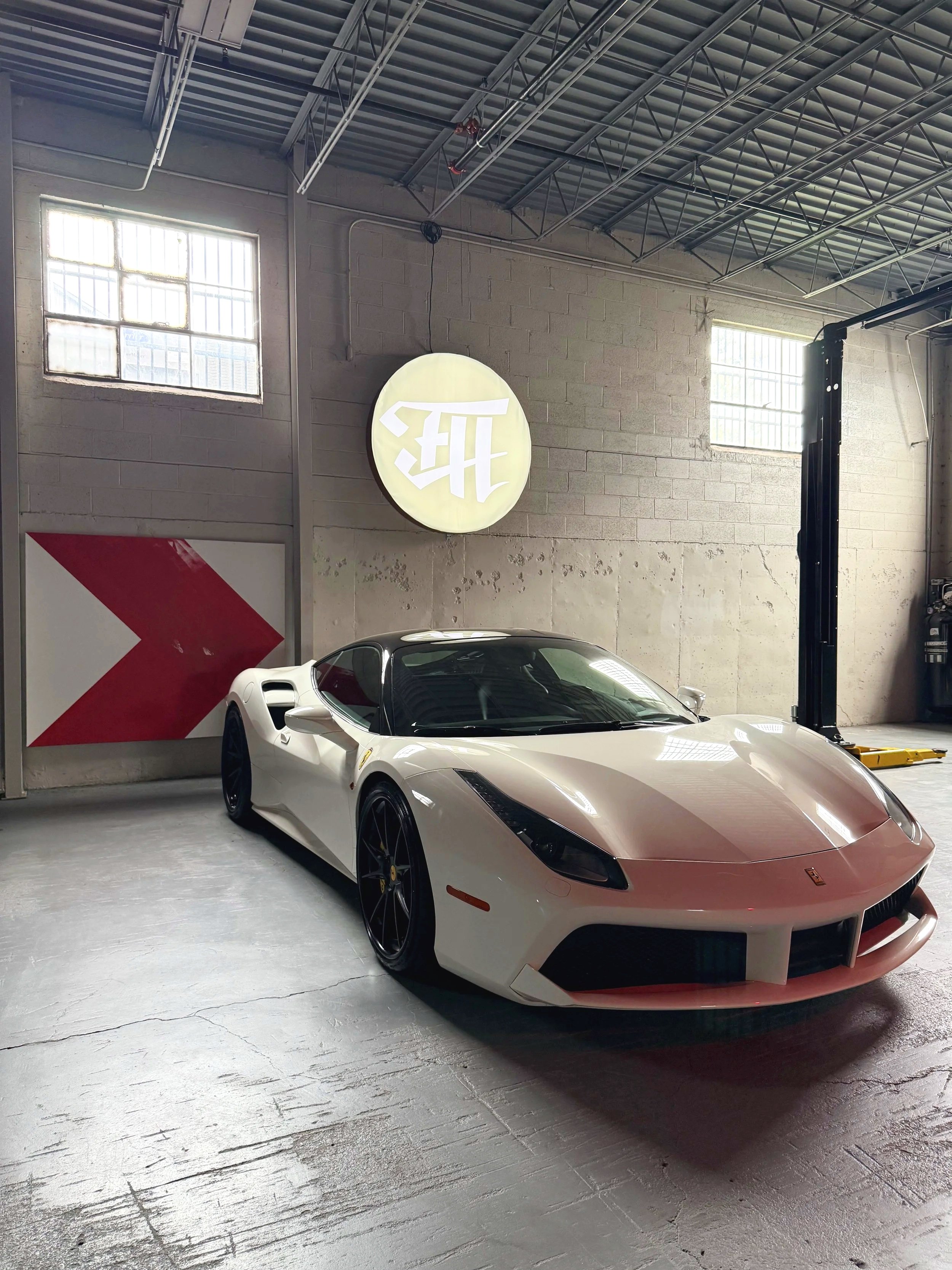 A white Ferrari sports car with black wheels parked inside a garage with concrete walls and large windows.