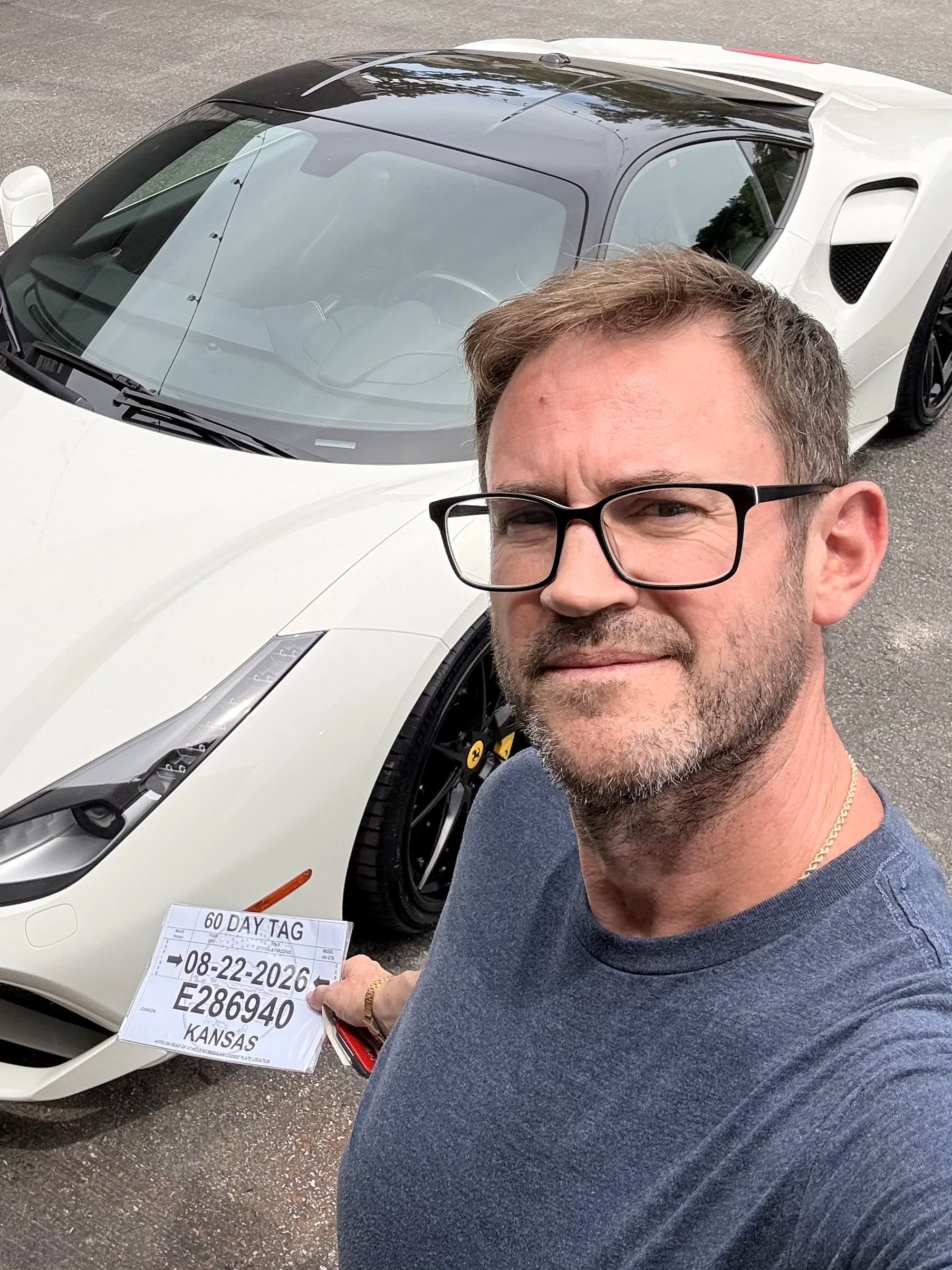 A man with glasses, gray hair, and a beard taking a selfie in front of a white sports car with a black roof. The man is holding a paper indicating a 60-day tag, with a date of 08-22-2026, and a license plate number. The man is wearing a dark t-shirt and a chain necklace.