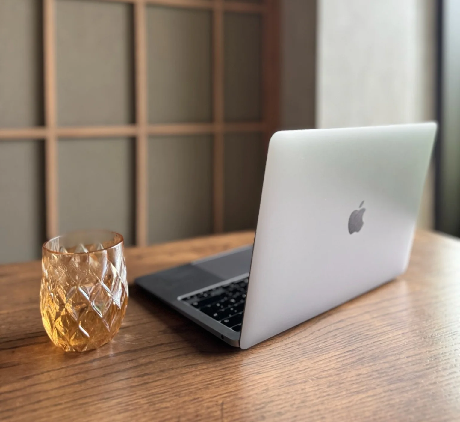 Ein MacBook auf einem Holztisch neben einem braunen Glas. Hintergrund mit einer Holzregalwand.
