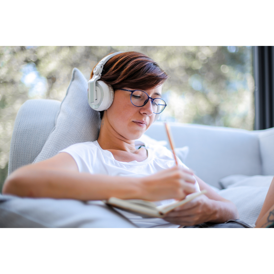 Frau mit Brille und Kopfhörern sitzt auf einem hellen Sofa und schreibt in ein Notizbuch.