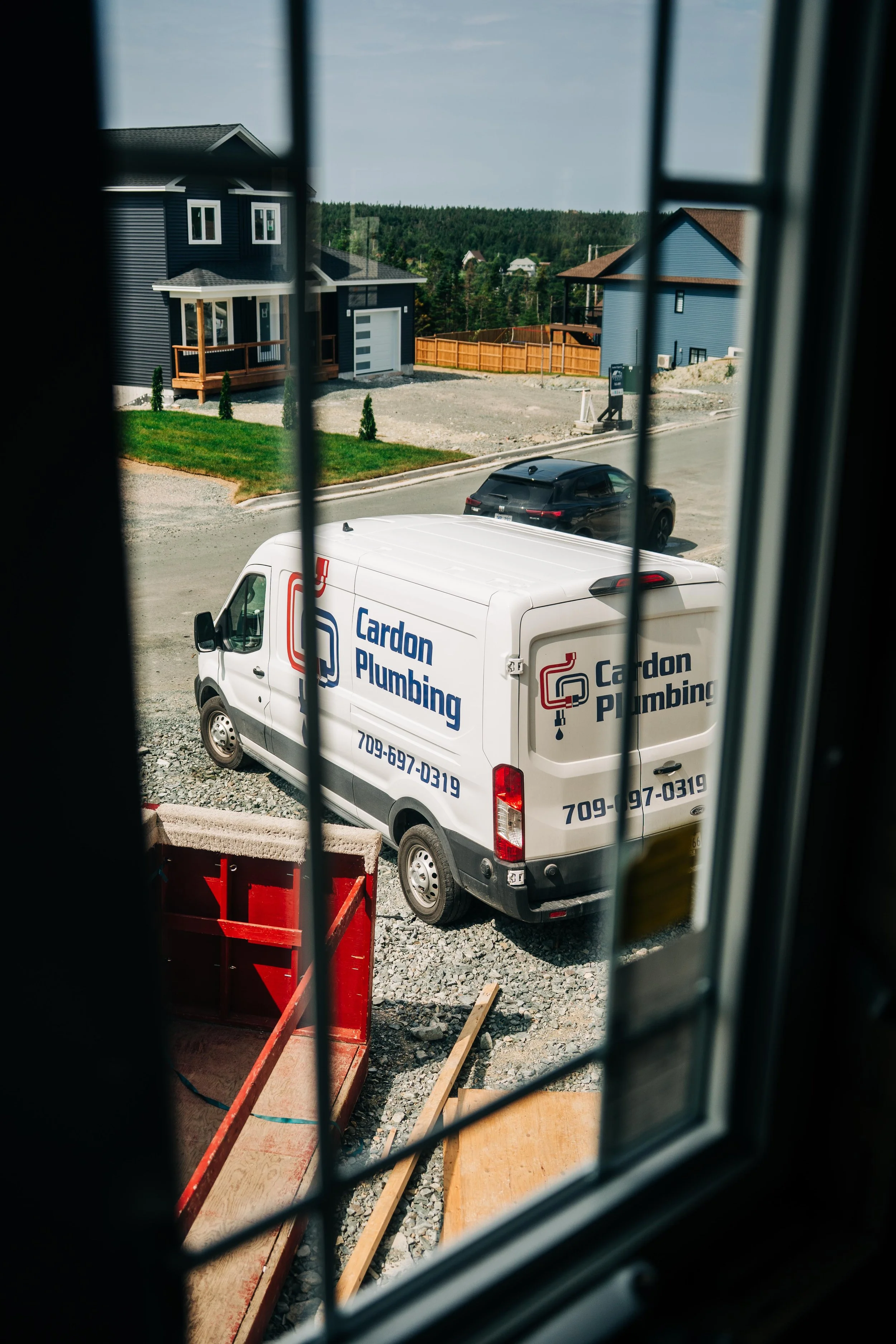 Road construction site seen through a window with black bars. There are construction materials, a white service van with 'Cardon Plumbing' written on it, a black car, and houses in the background on a sunny day.