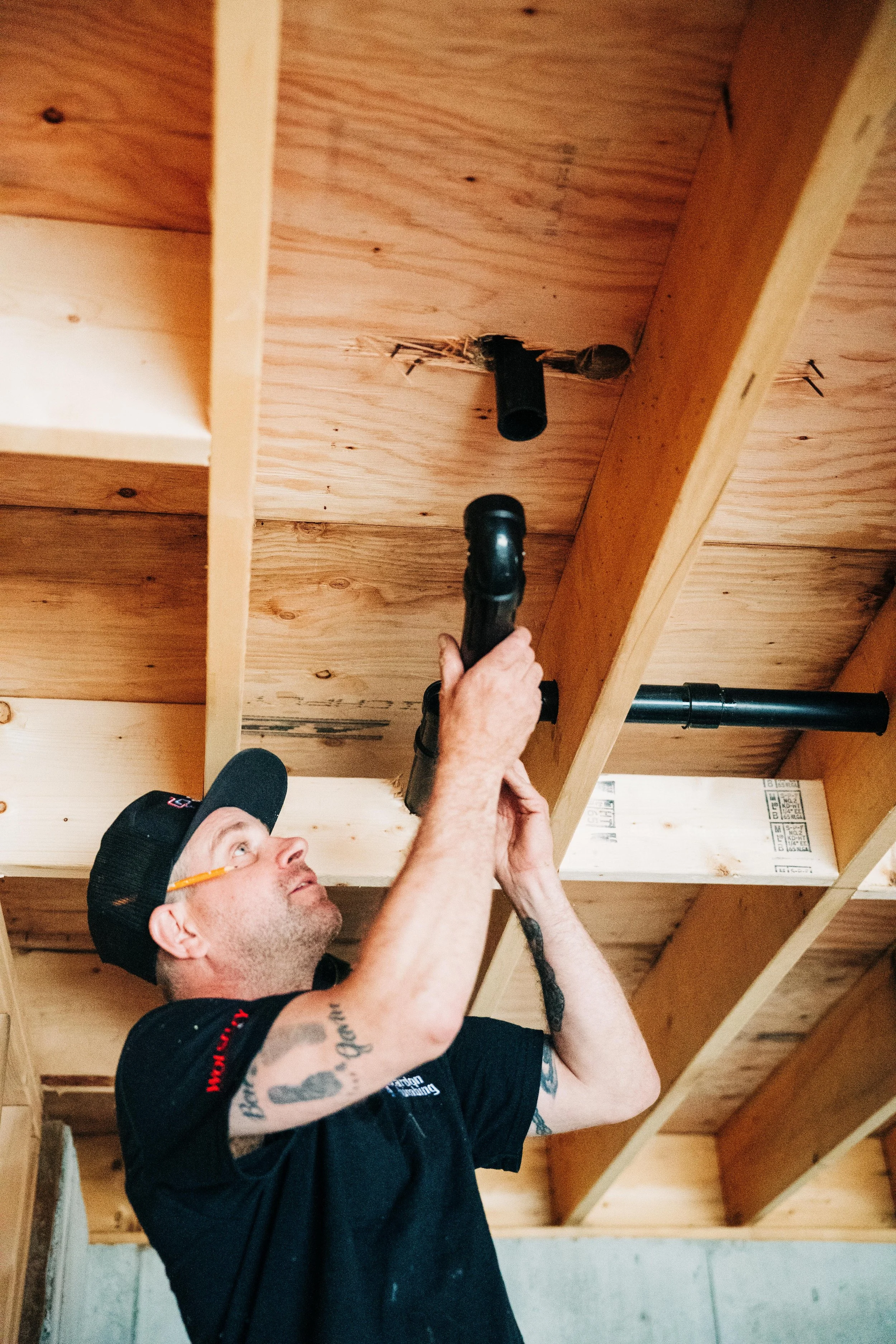 A man wearing a black cap, black T-shirt, glasses, and tattoos on his arm is holding a drill and working on the wooden ceiling of a room construction site.
