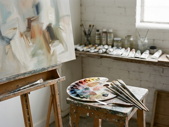 An artist's studio with an abstract painting on an easel, a paint palette, brushes, and tubes of paint on a table, with jars of brushes and paint on a windowsill in the background.