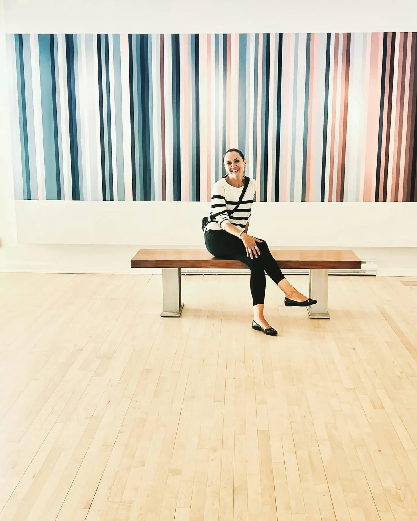 A woman sitting on a wooden bench in front of a colorful striped abstract painting, smiling at the camera.