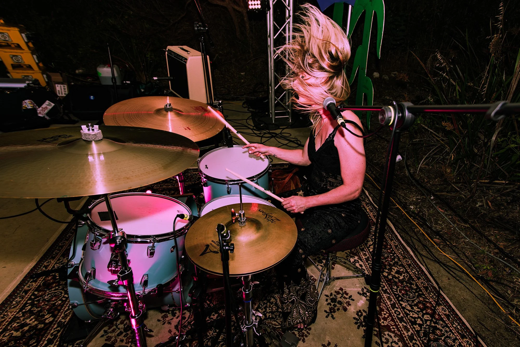 A woman with blonde hair playing the drums on a stage at night, with musical equipment and plants in the background.